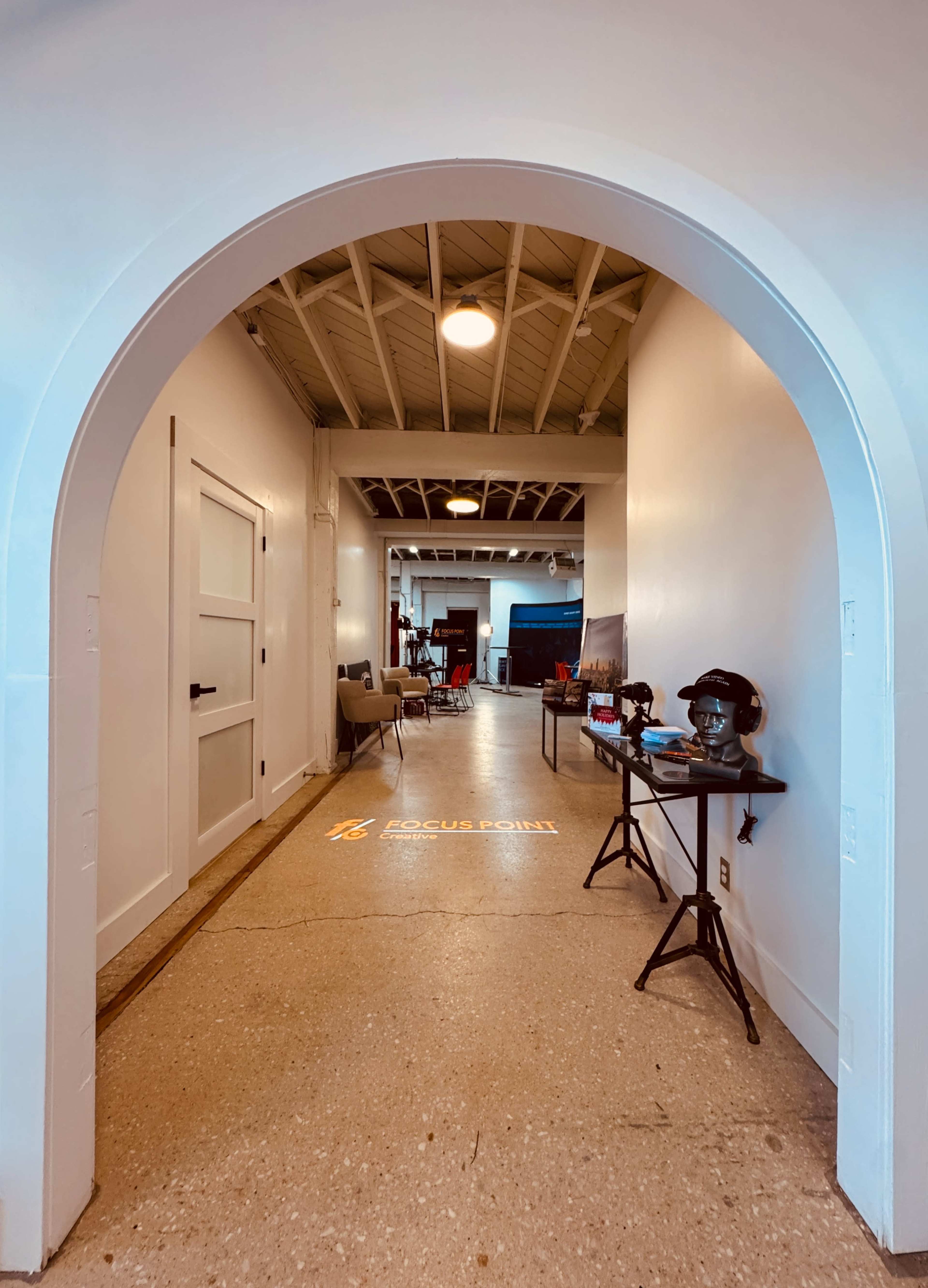The image shows a well-lit hallway framed by an arch, leading to a spacious area with various furniture items and artwork along the walls.