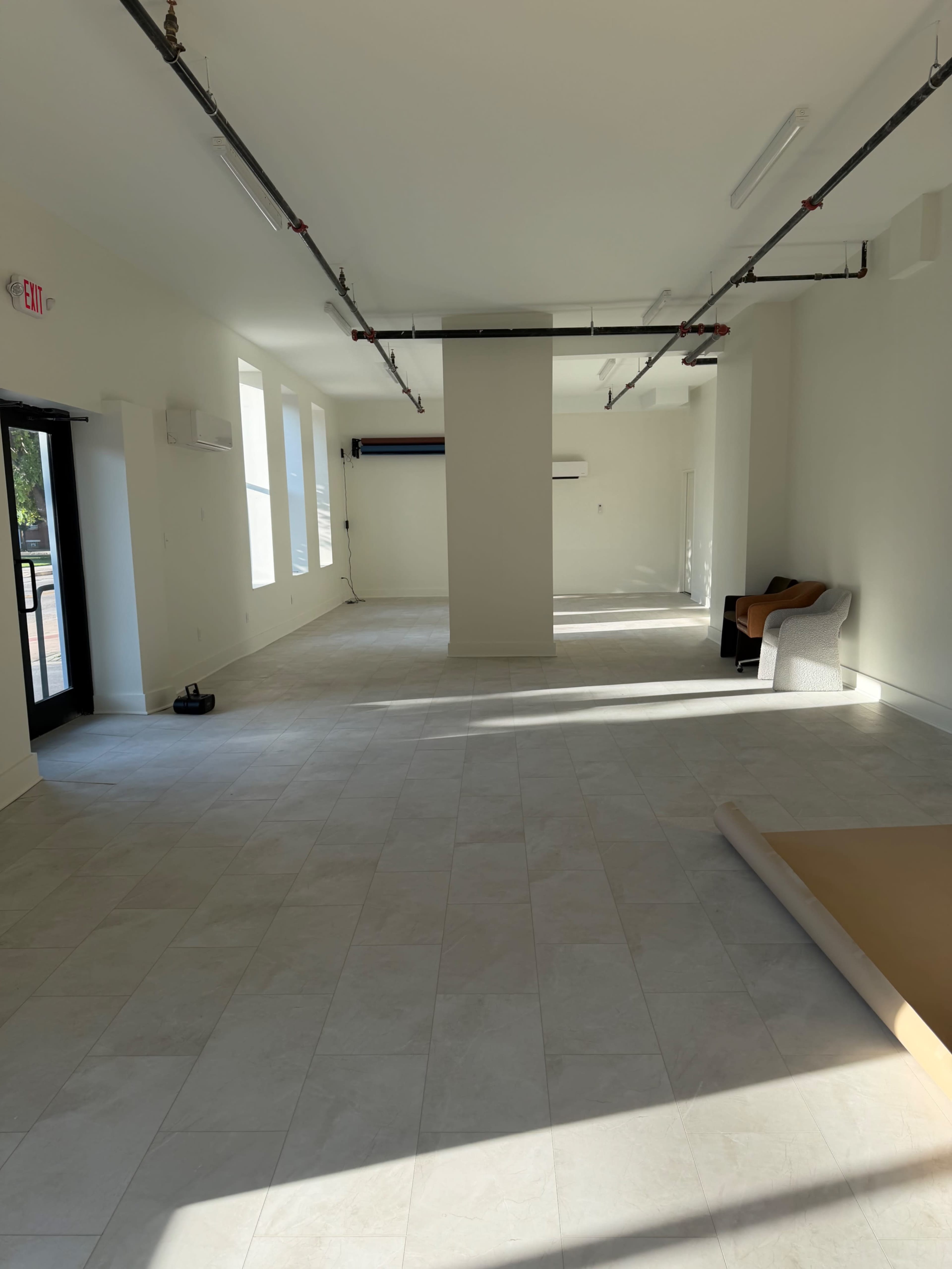 The image shows an empty, well-lit interior space with neutral walls, large windows, and a few pieces of furniture placed against the wall.