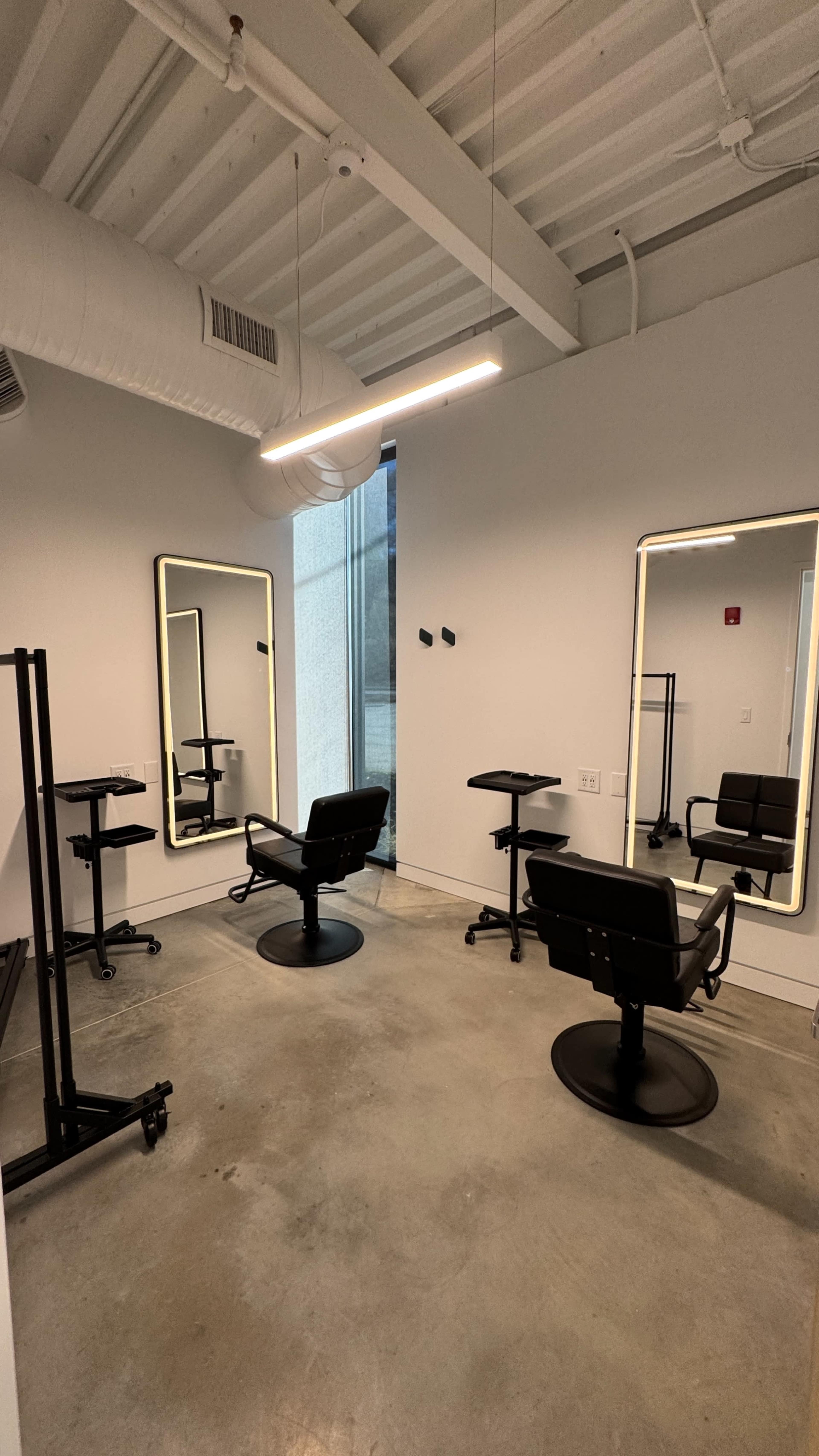 The image shows a minimalist salon interior featuring two mirrors, two styling chairs, and a couple of mobile carts arranged in a well-lit space.