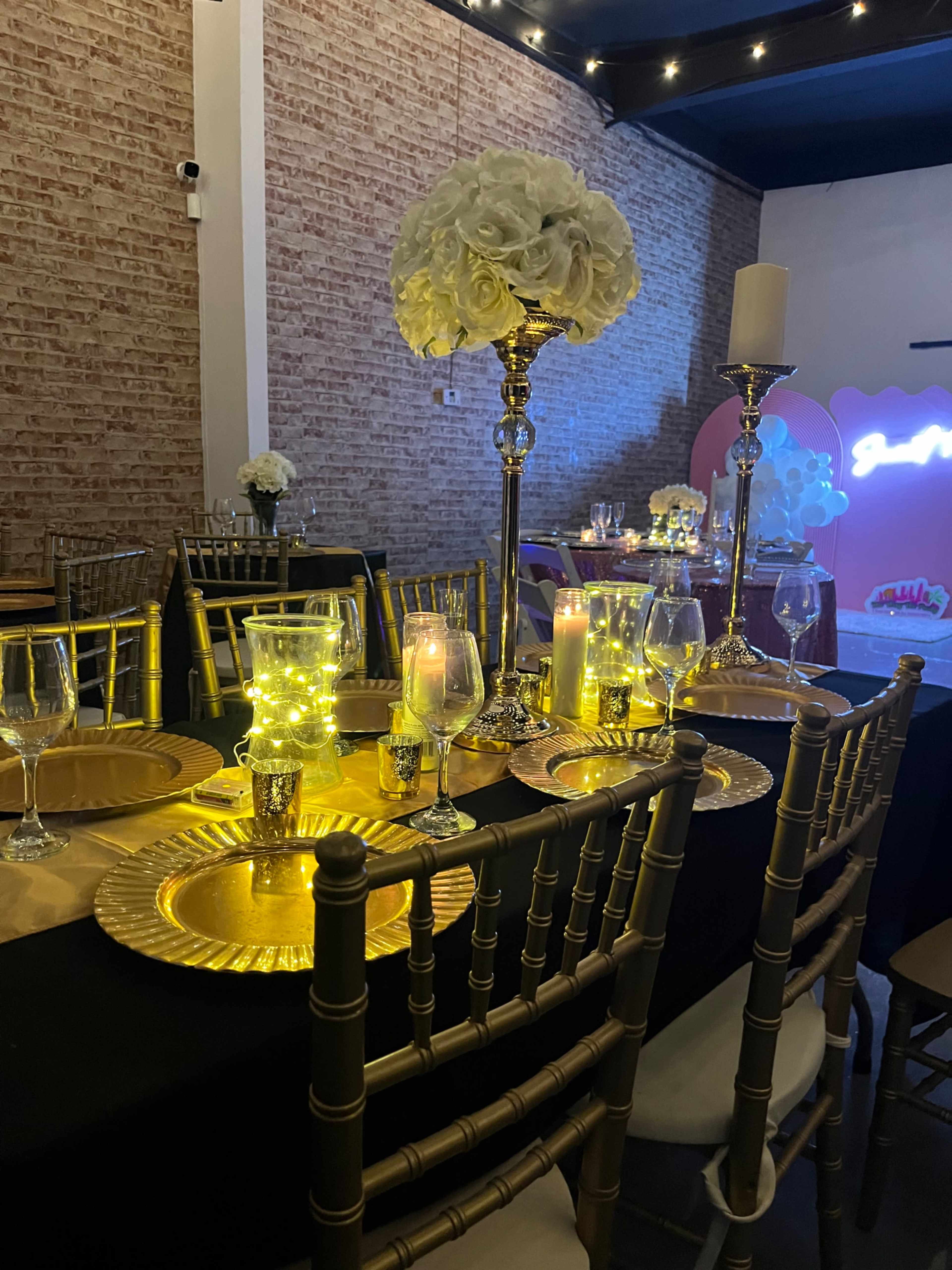 The image shows a banquet hall with elegantly set tables, featuring gold-colored plates, glassware, and floral centerpieces illuminated by soft lights.