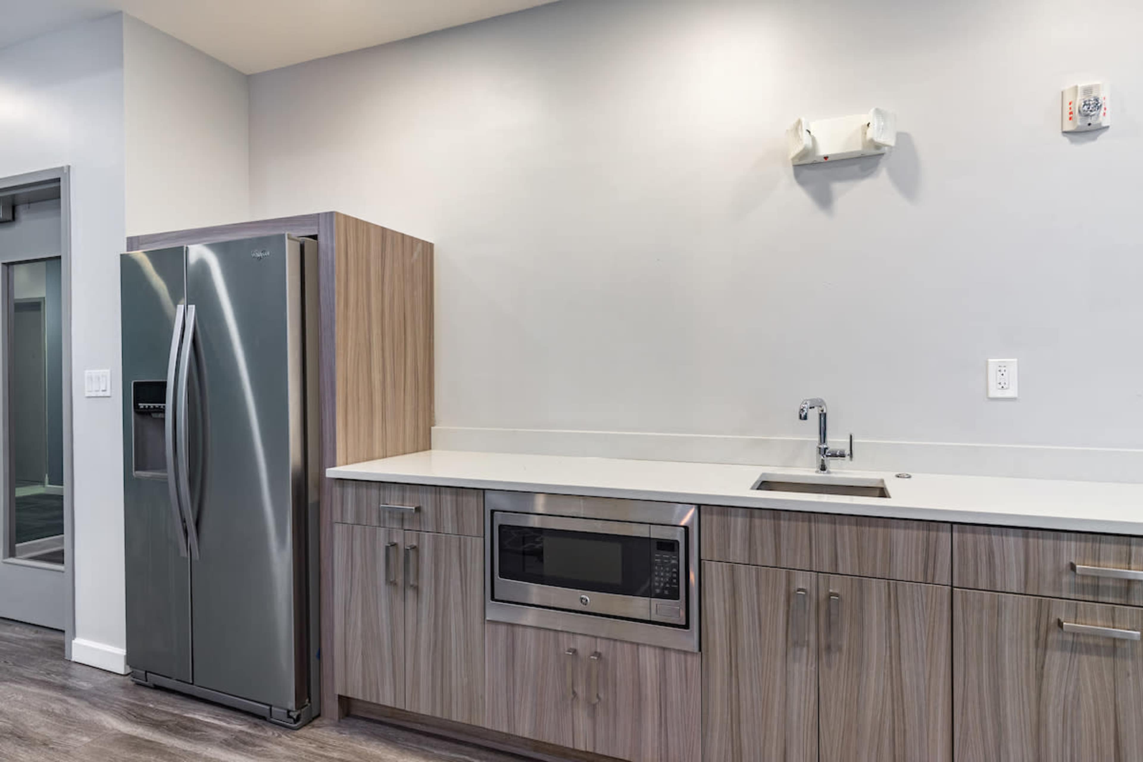 A modern kitchen with stainless steel appliances, including a refrigerator and microwave, against a light-colored wall and wooden cabinetry.