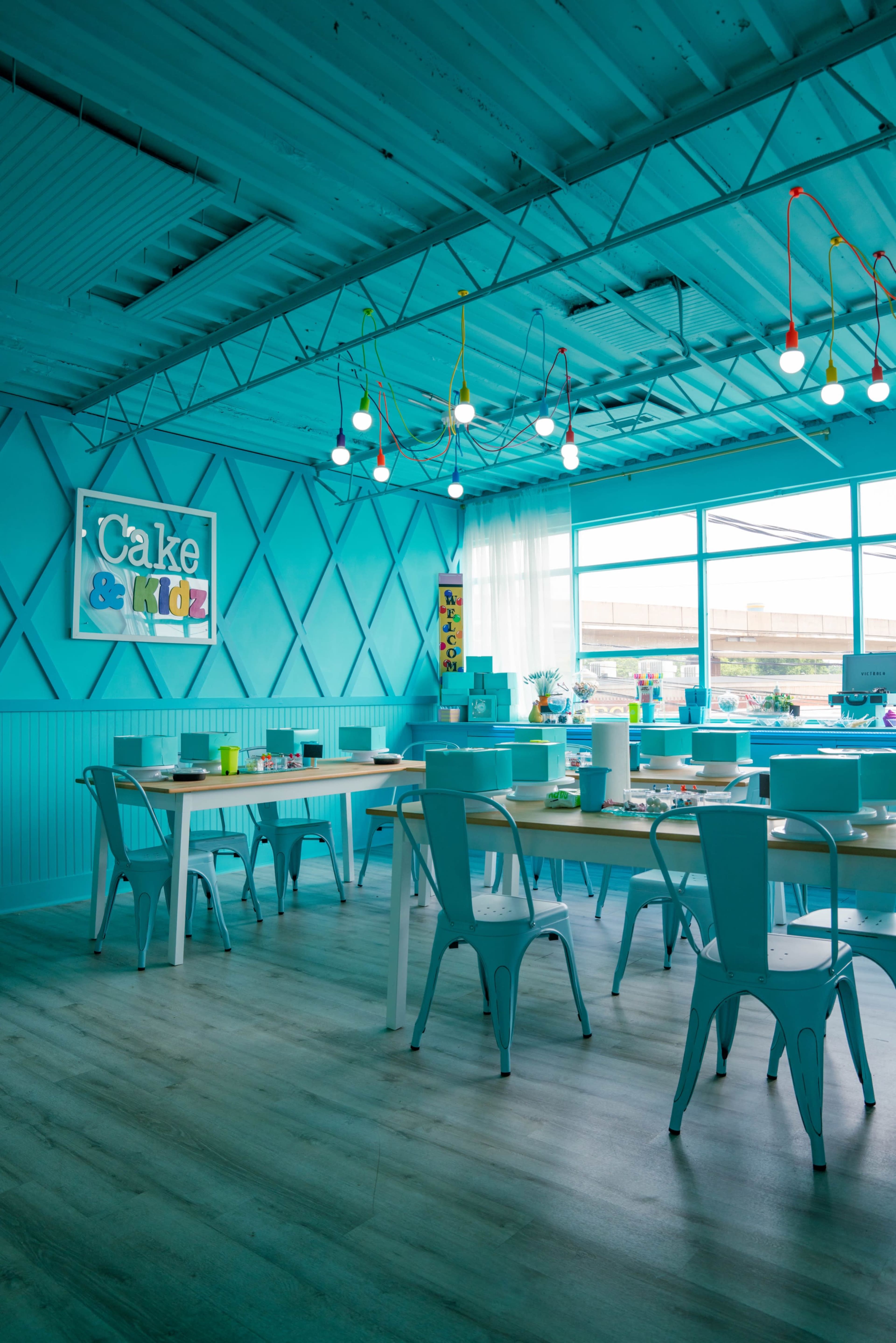 A brightly colored turquoise room with tables and chairs arranged for a baking or craft activity, featuring a sign that reads "Cake & Kids" on the wall.