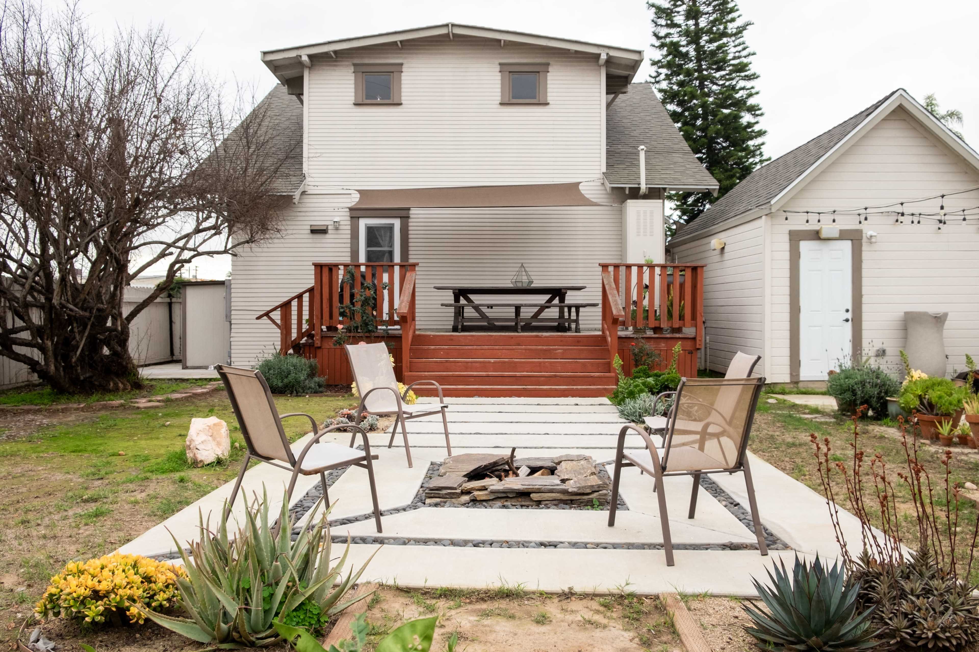 A two-story house with a wooden deck and a seating area surrounding a stone fire pit is situated in a landscaped yard.