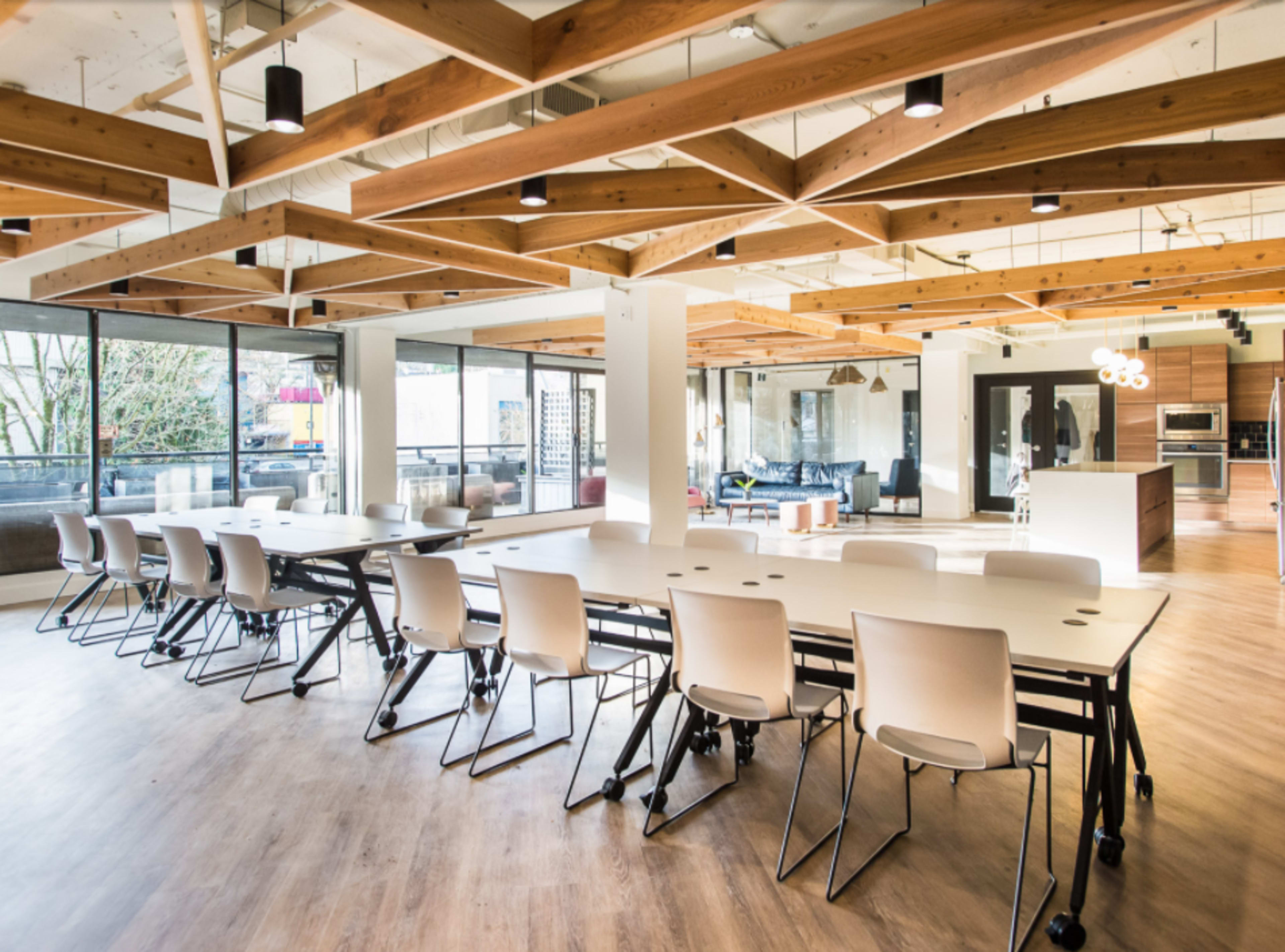 A modern office space featuring a large table surrounded by chairs, with wooden beams and large windows allowing natural light.