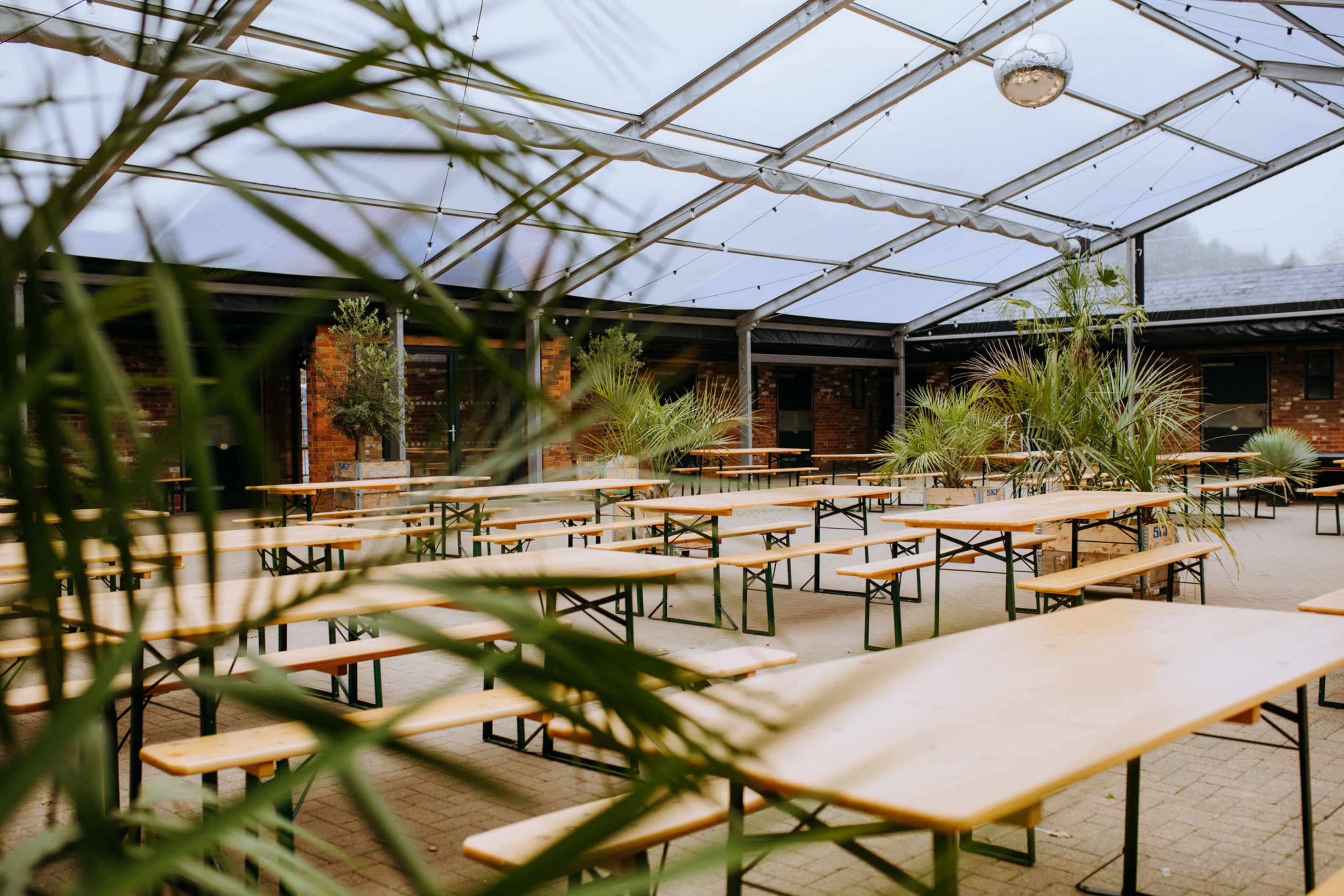 An outdoor event space features numerous wooden tables arranged under a transparent roof, surrounded by potted plants.