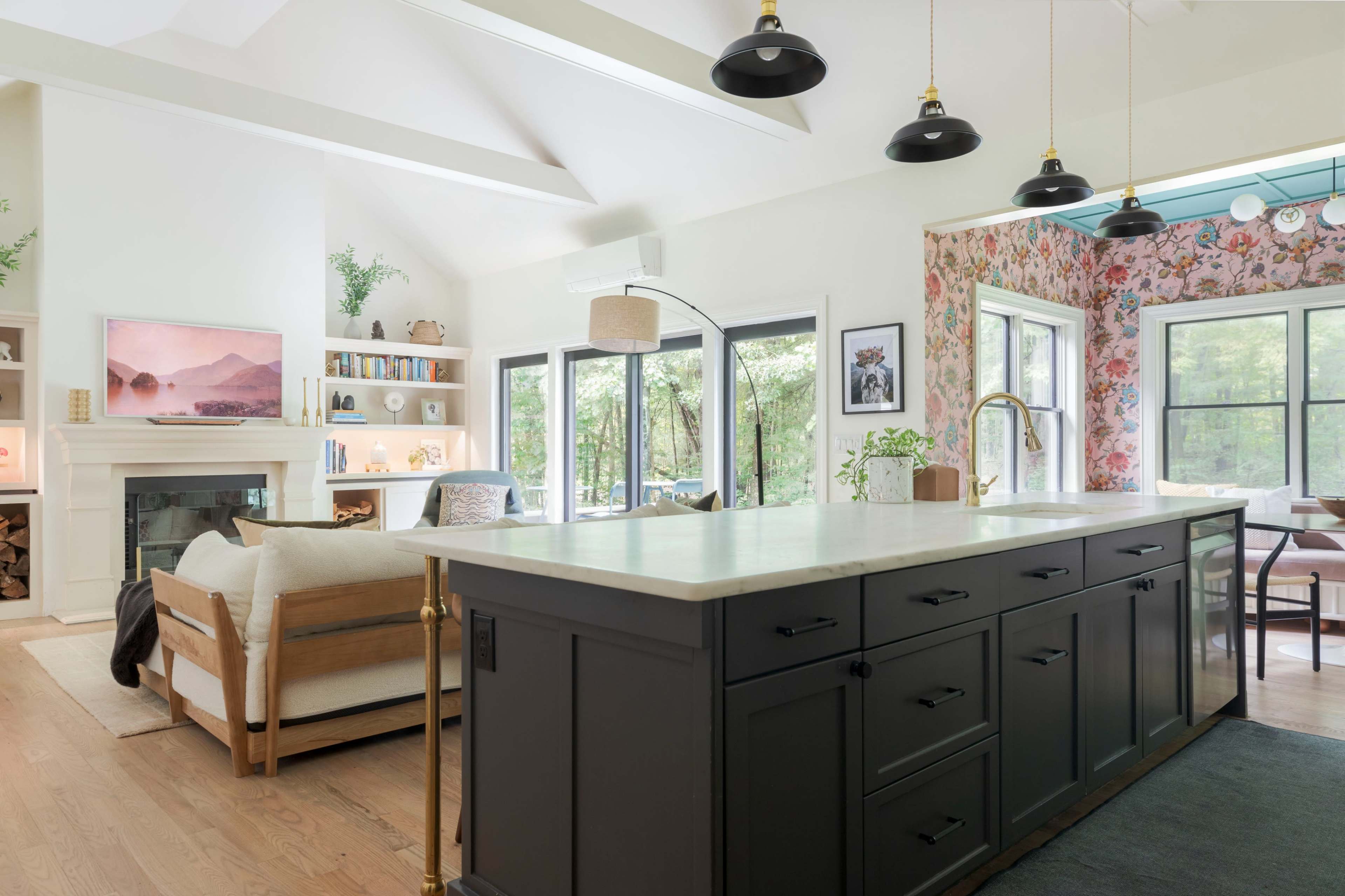 The image shows a modern kitchen with a large island, black cabinetry, and a living area featuring a sofa, bookshelves, and floral wallpaper.