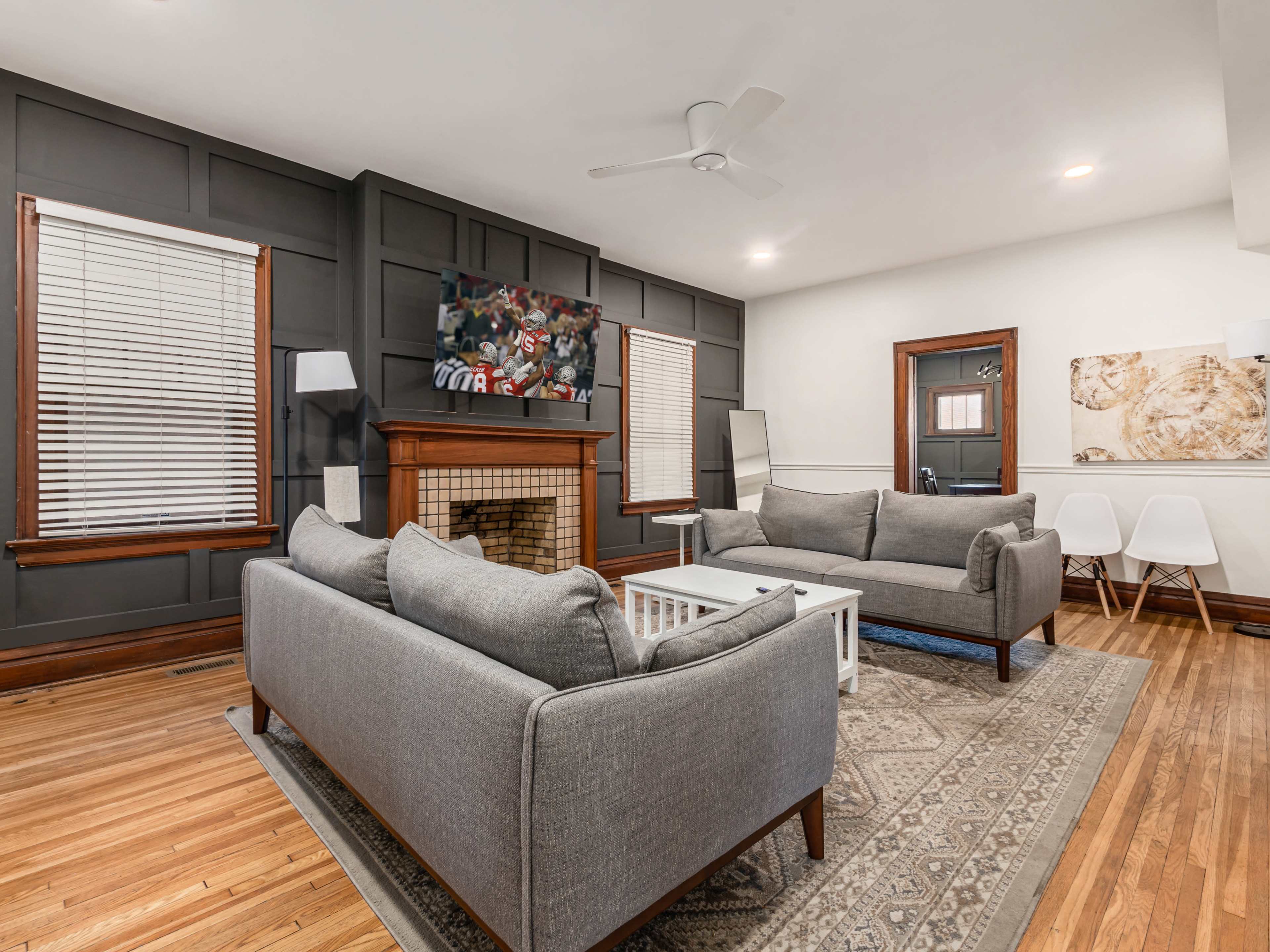 The living room features two gray sofas facing a wooden fireplace, a television mounted on the wall, and a coffee table on a patterned rug.