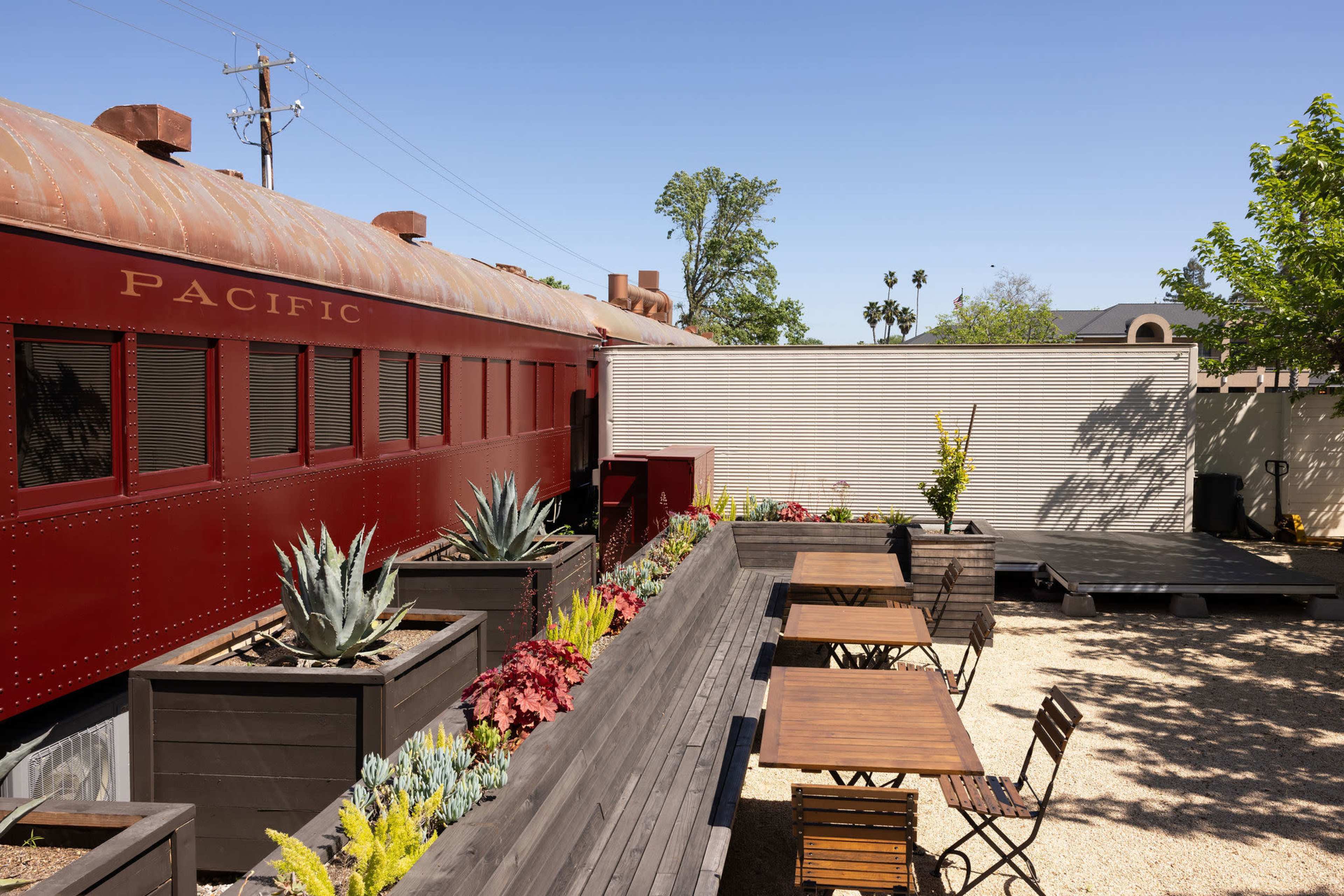A red train car is positioned beside a garden area with wooden tables and chairs, surrounded by succulents and desert plants.
