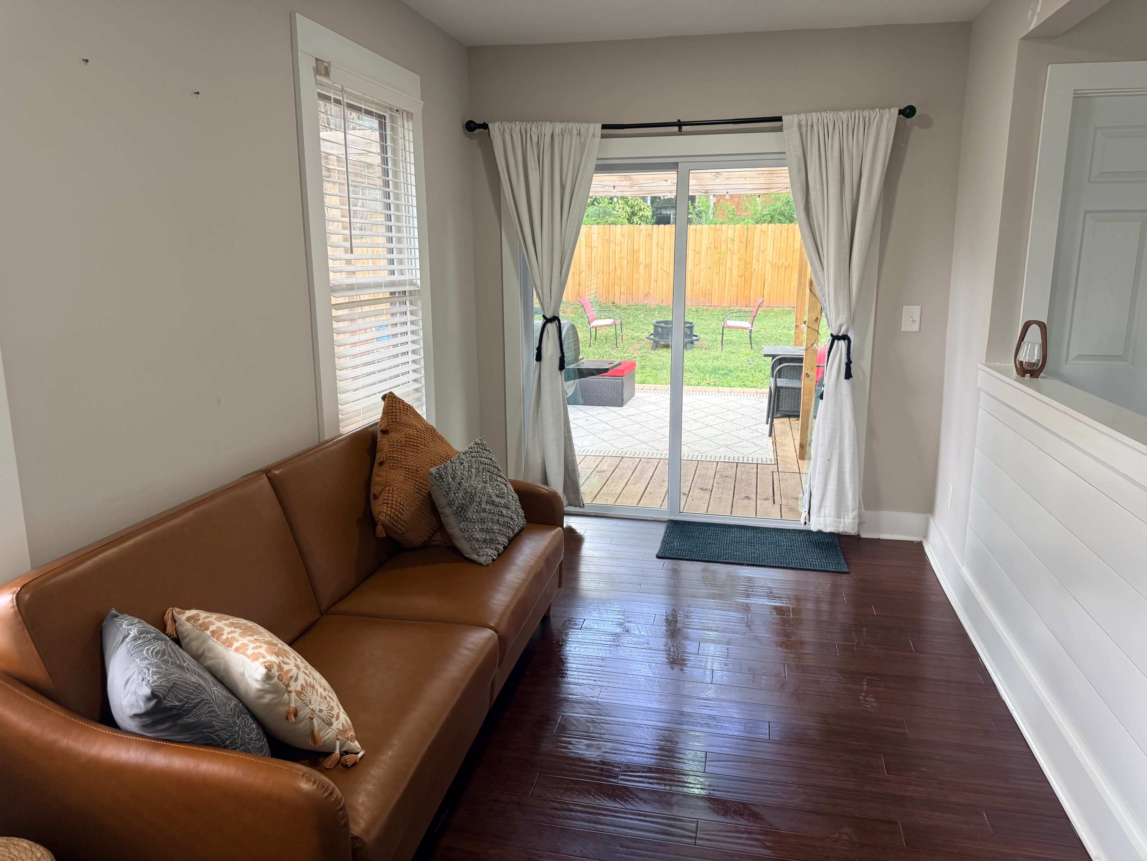 A brown leather couch is positioned in a well-lit living area, facing a sliding glass door that opens to a backyard patio with outdoor seating.