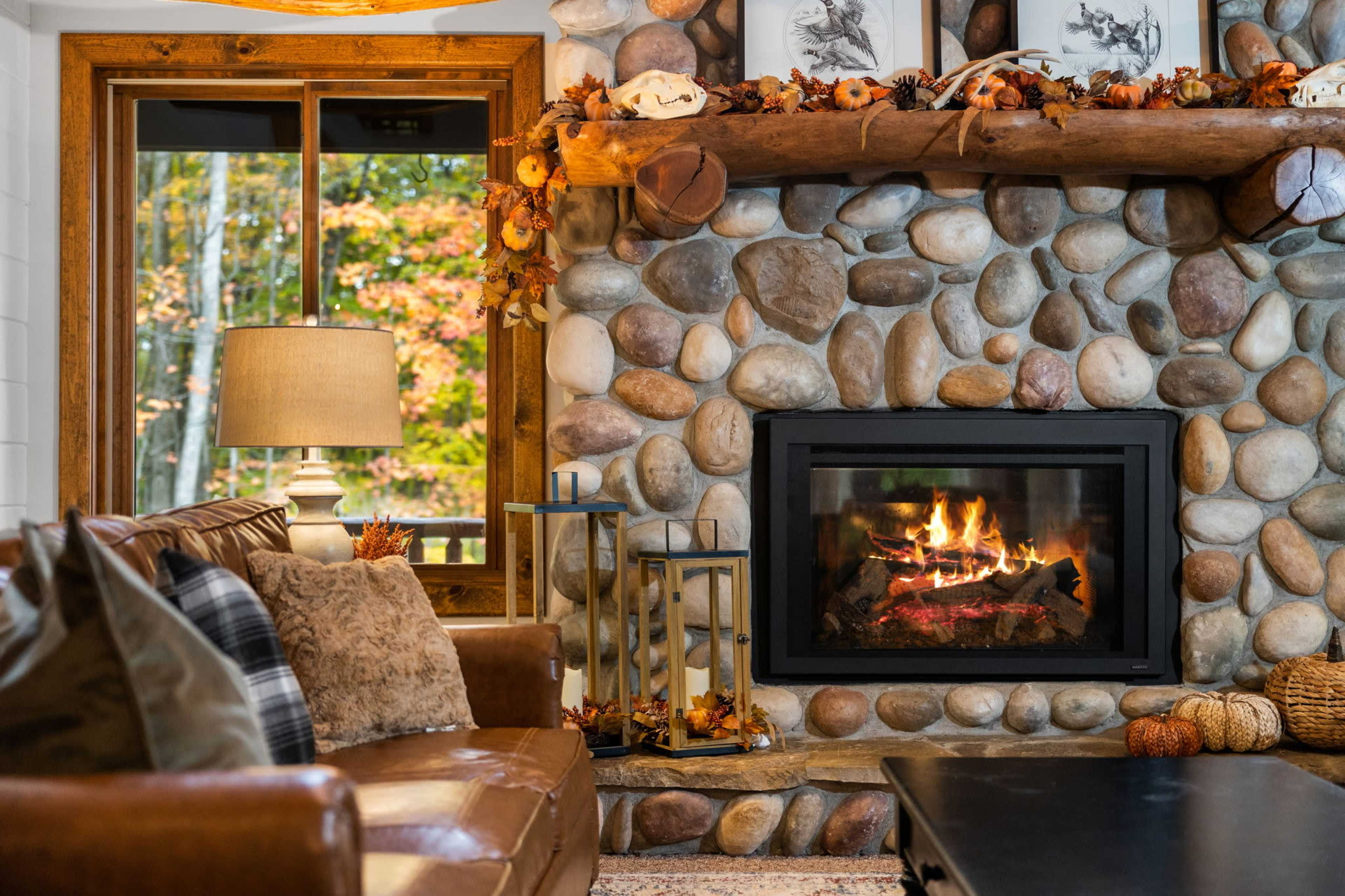 A cozy living room features a stone fireplace with a fire burning, surrounded by a leather sofa and decorative autumn-themed elements.