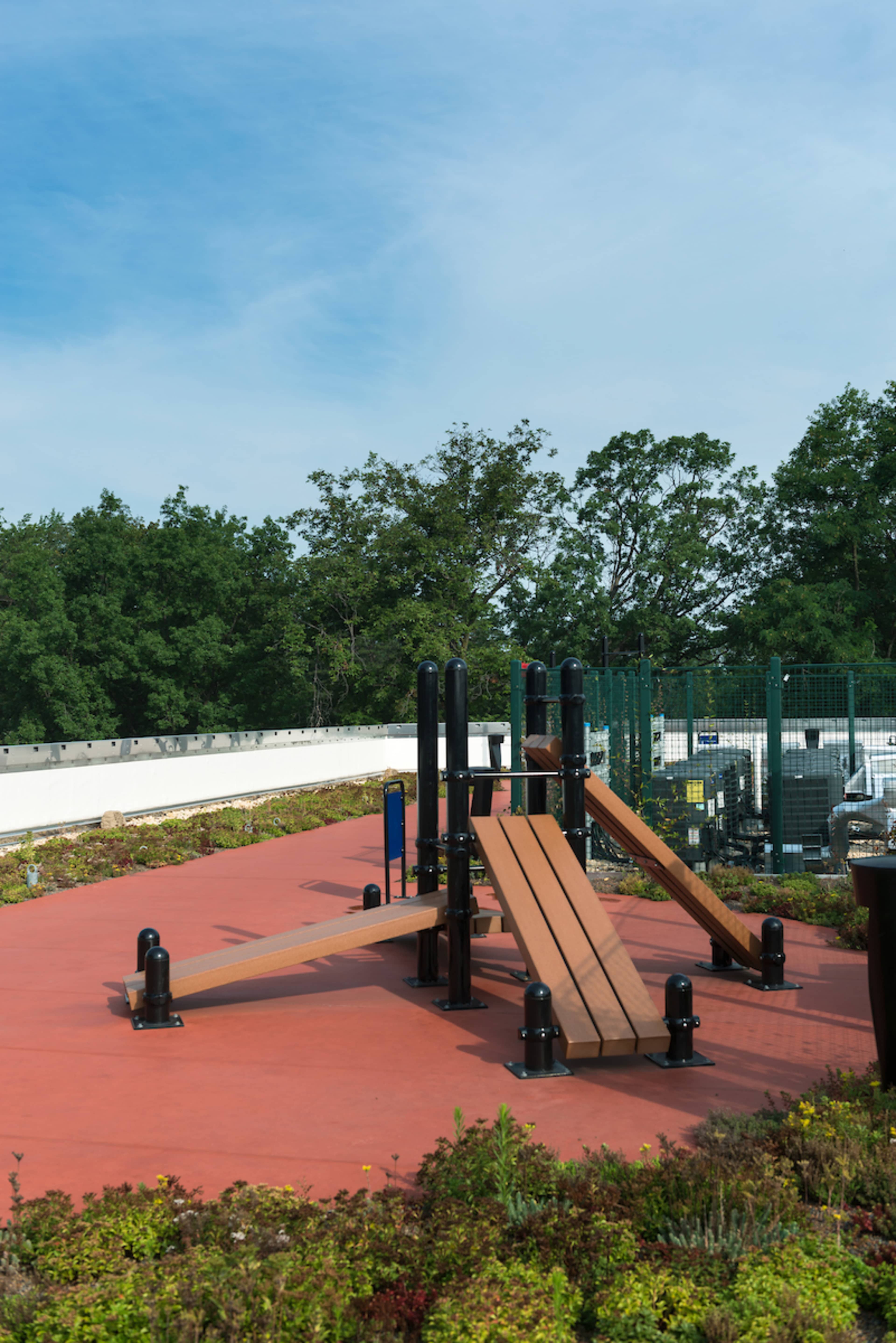 An outdoor fitness area with exercise equipment, surrounded by greenery and a fence in the background.