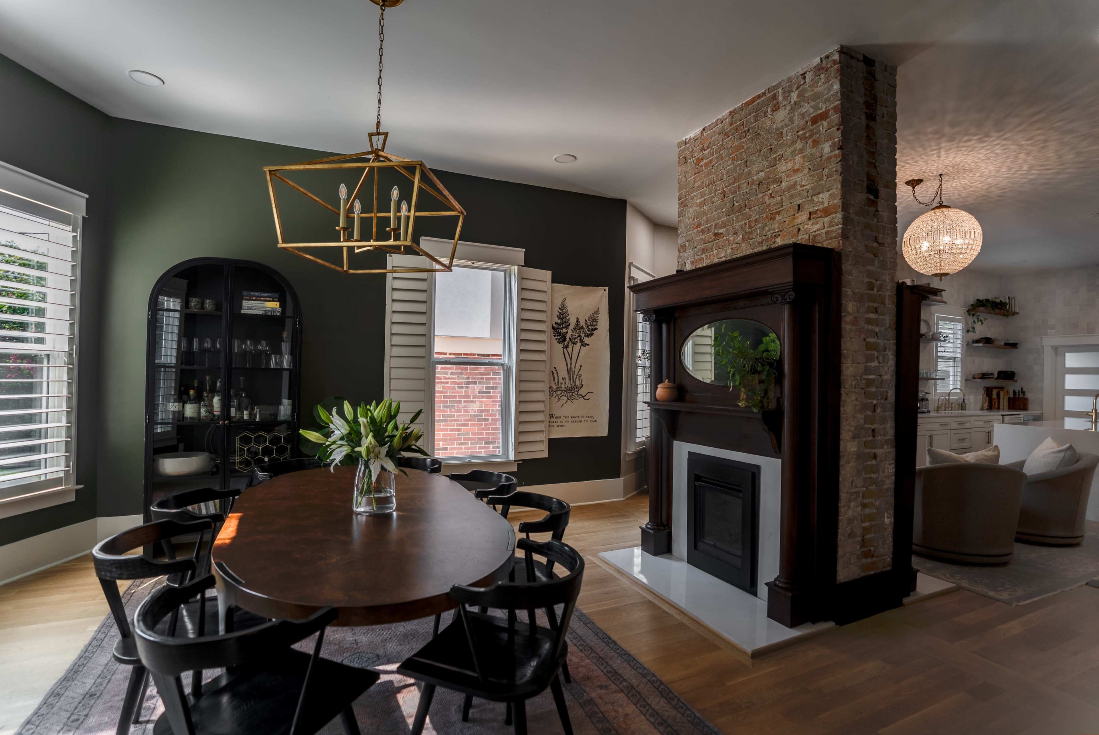 The image shows a dining area with a circular wooden table surrounded by black chairs, a modern light fixture hanging above, and a brick fireplace adjacent to a cozy living space.