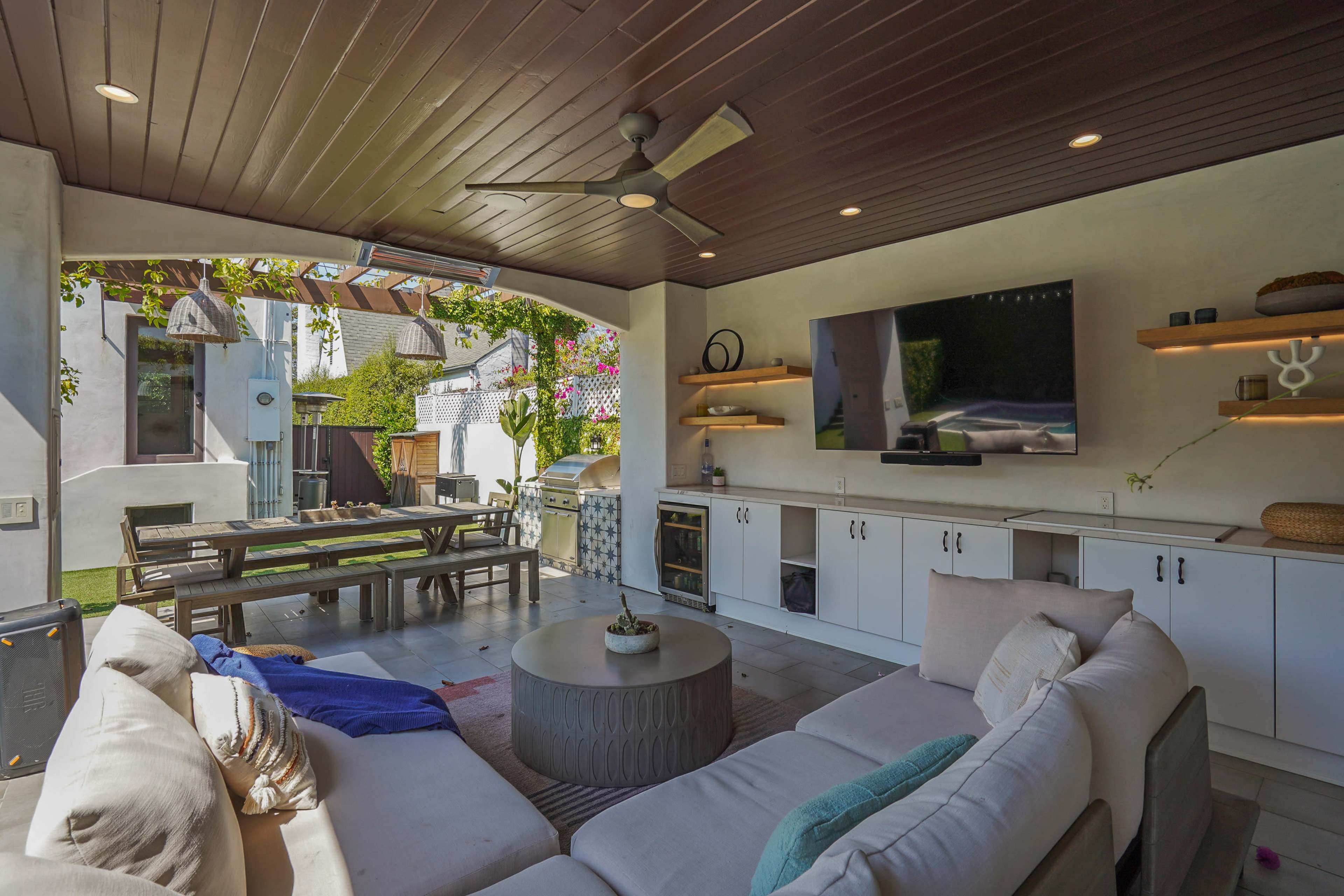 The image shows a covered outdoor seating area featuring a large sectional sofa, a round coffee table, and a flat-screen television mounted on the wall.