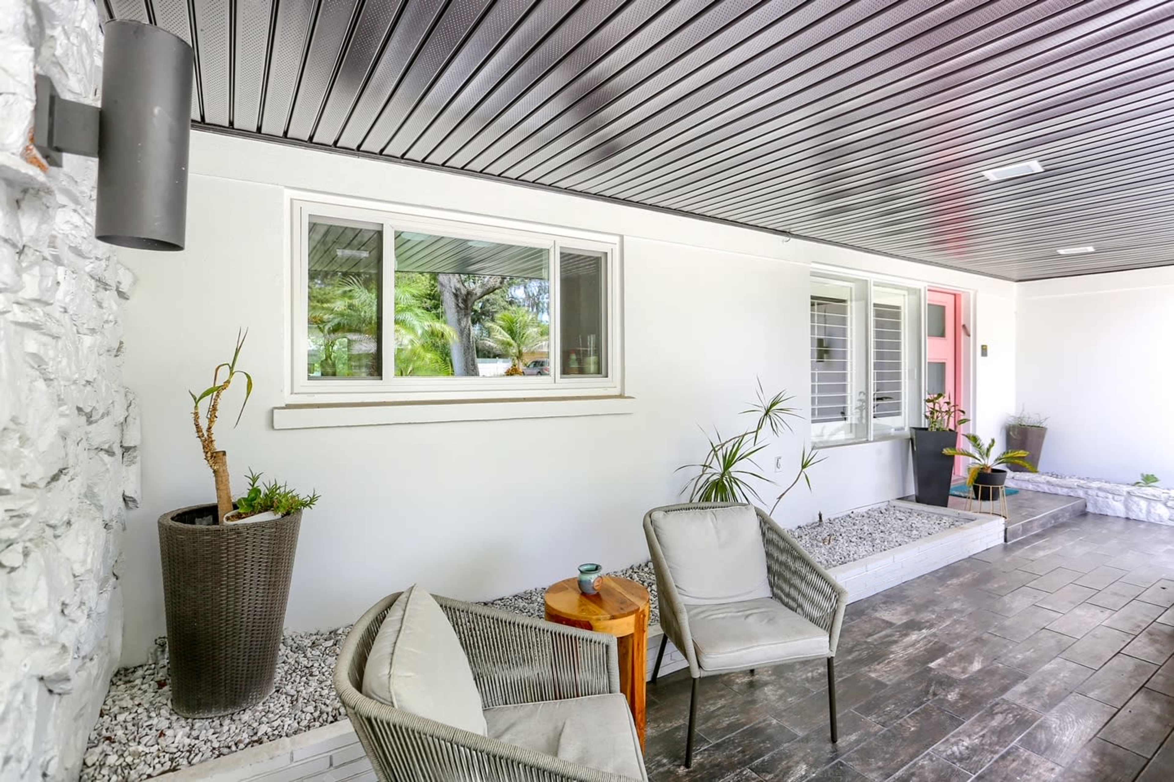 A modern porch area with two chairs, a small table, and potted plants along a stone pathway beside a white wall.