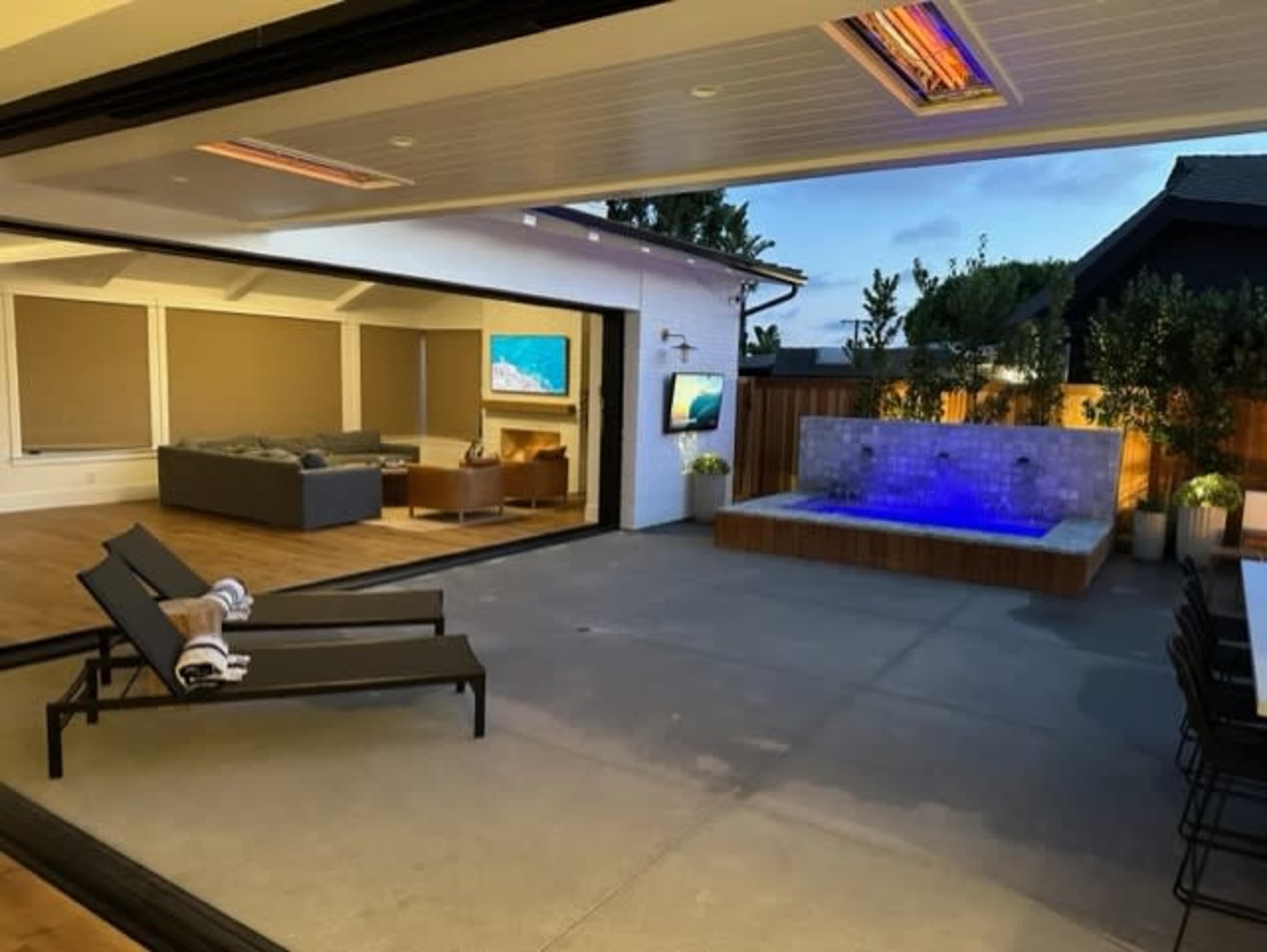 A modern outdoor space features a hot tub with blue illumination, a seating area with a gray couch, and large sliding glass doors that open to the interior.