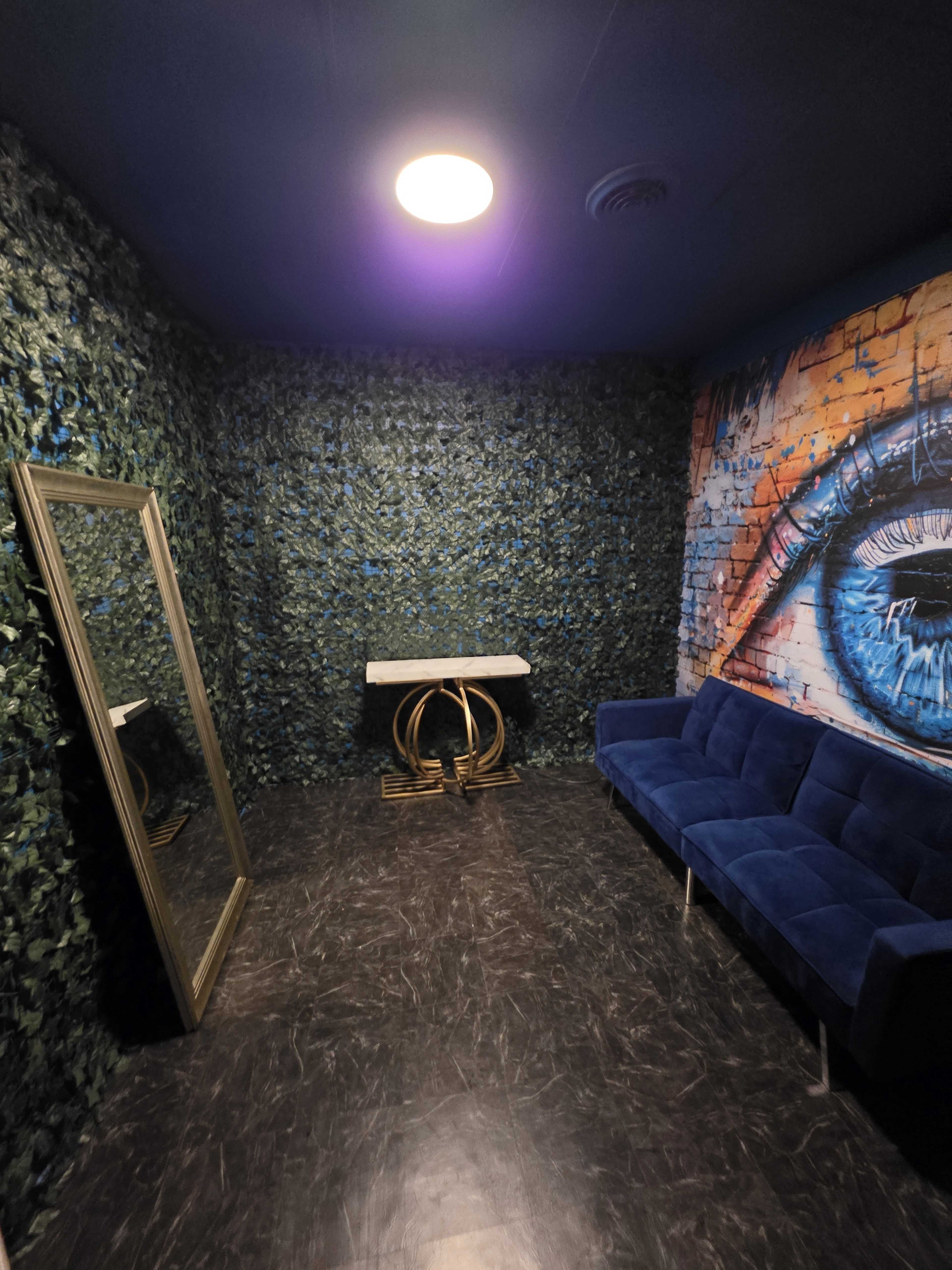 The image shows a small room with a dark floor, a blue velvet couch, a marble table, a large mirror, and green foliage wallpaper, accented by a mural of an eye on one wall.