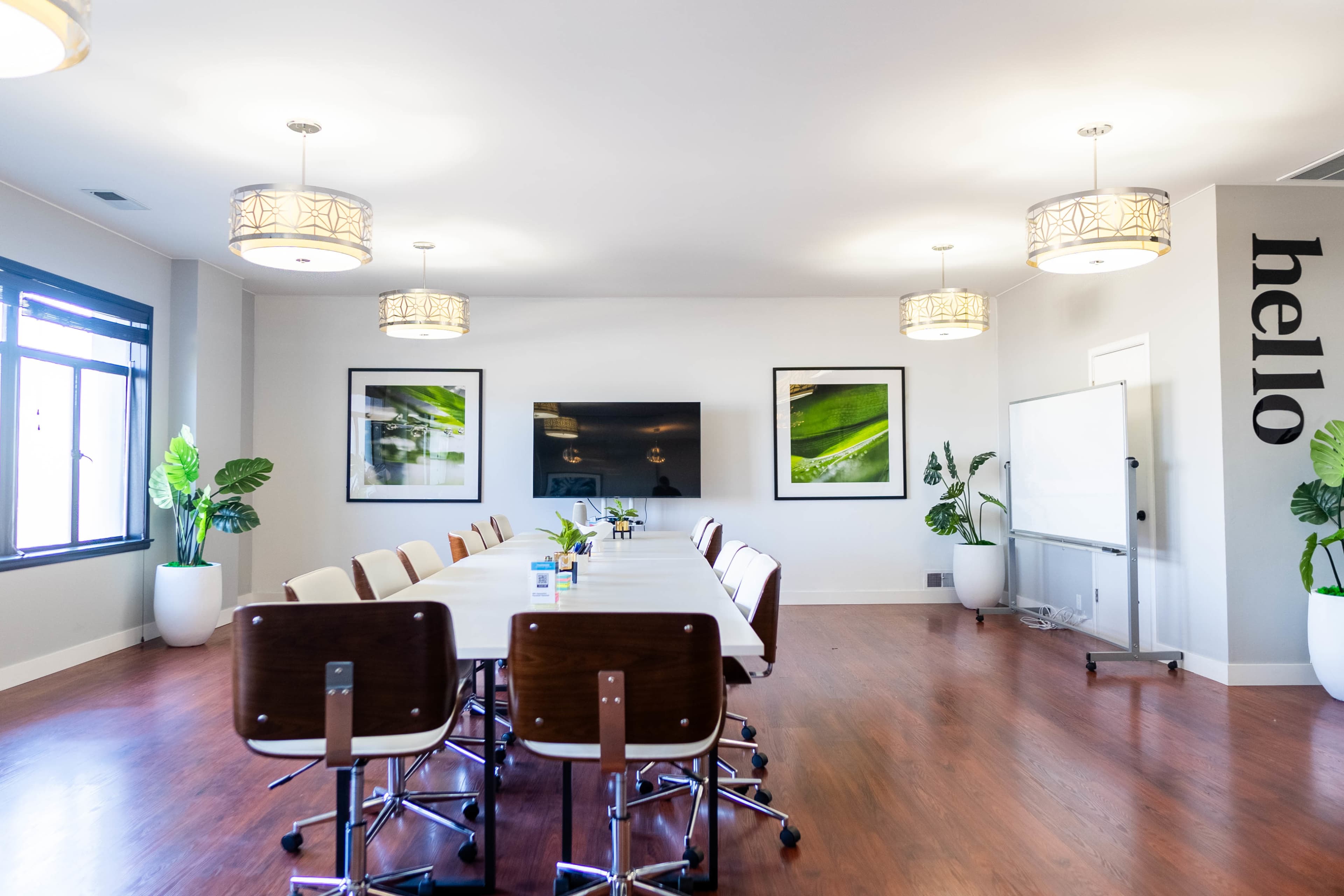 A modern conference room features a long table surrounded by chairs, with plants, artwork, and a large screen on the wall.