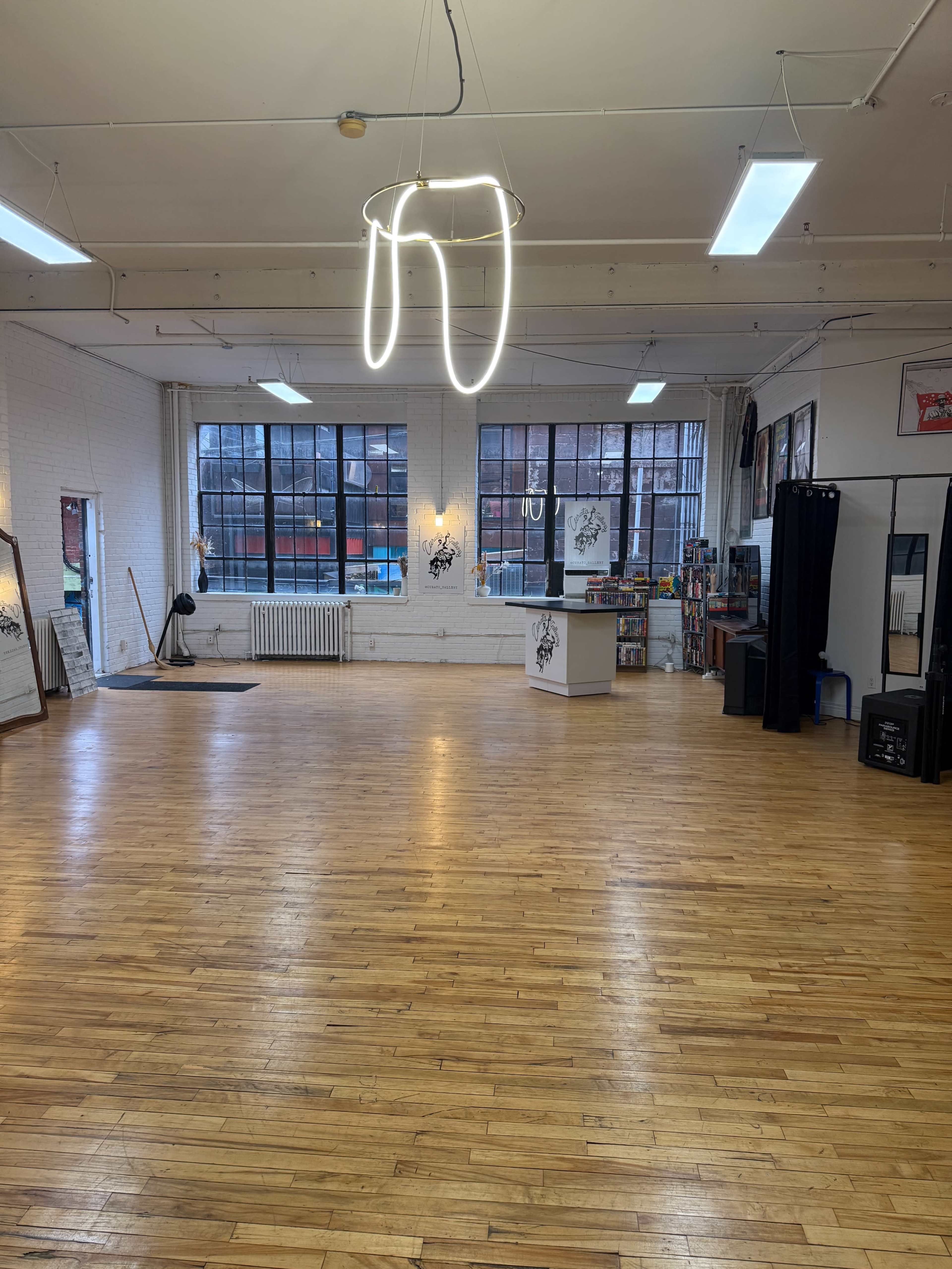 The image shows a spacious, well-lit studio with wooden flooring, large windows, and minimal furnishings, including a front desk and a mirror.