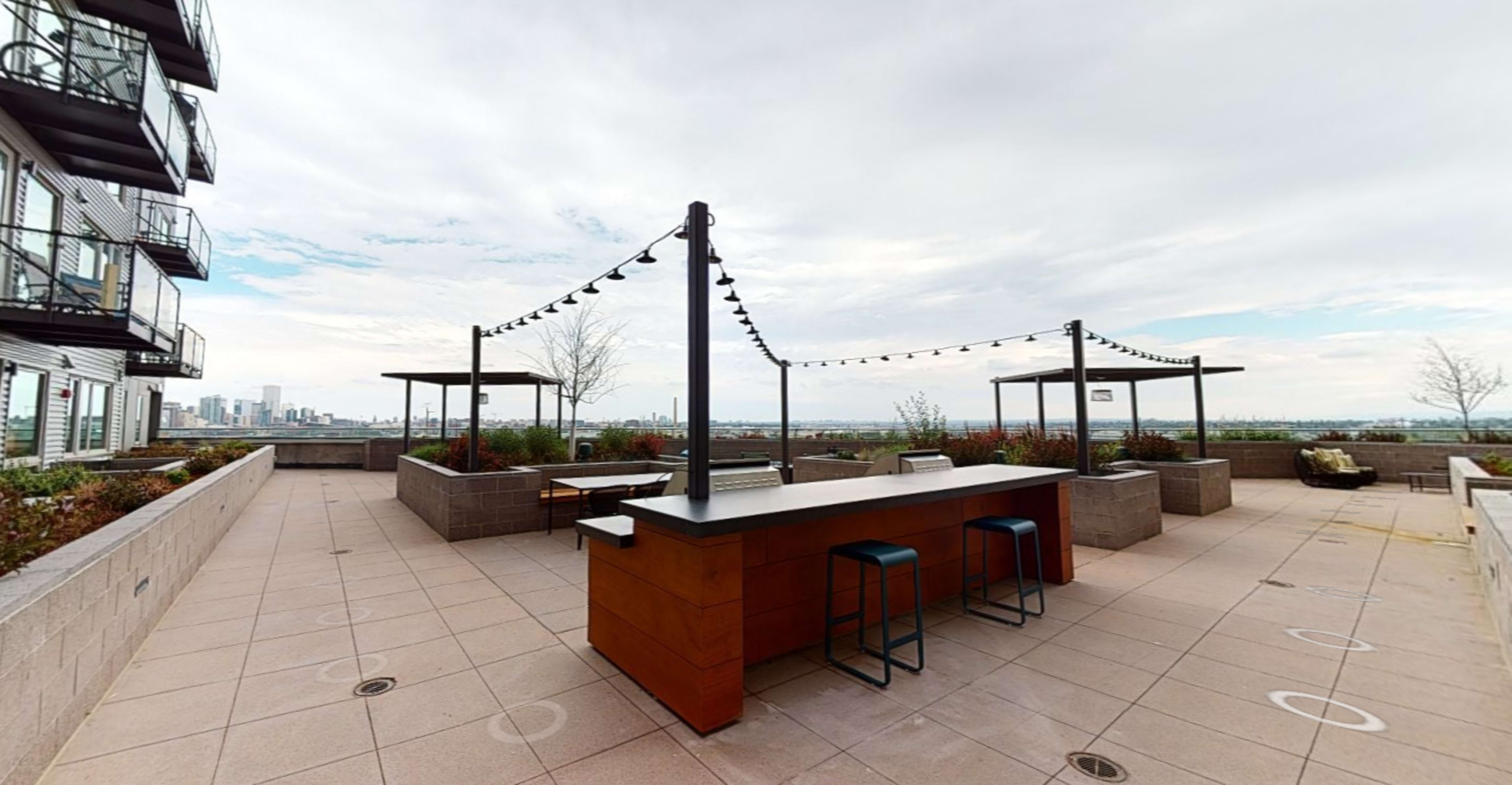A rooftop patio area featuring a long bar with stools, surrounded by overhead lights and greenery, with a view of a city skyline in the distance.