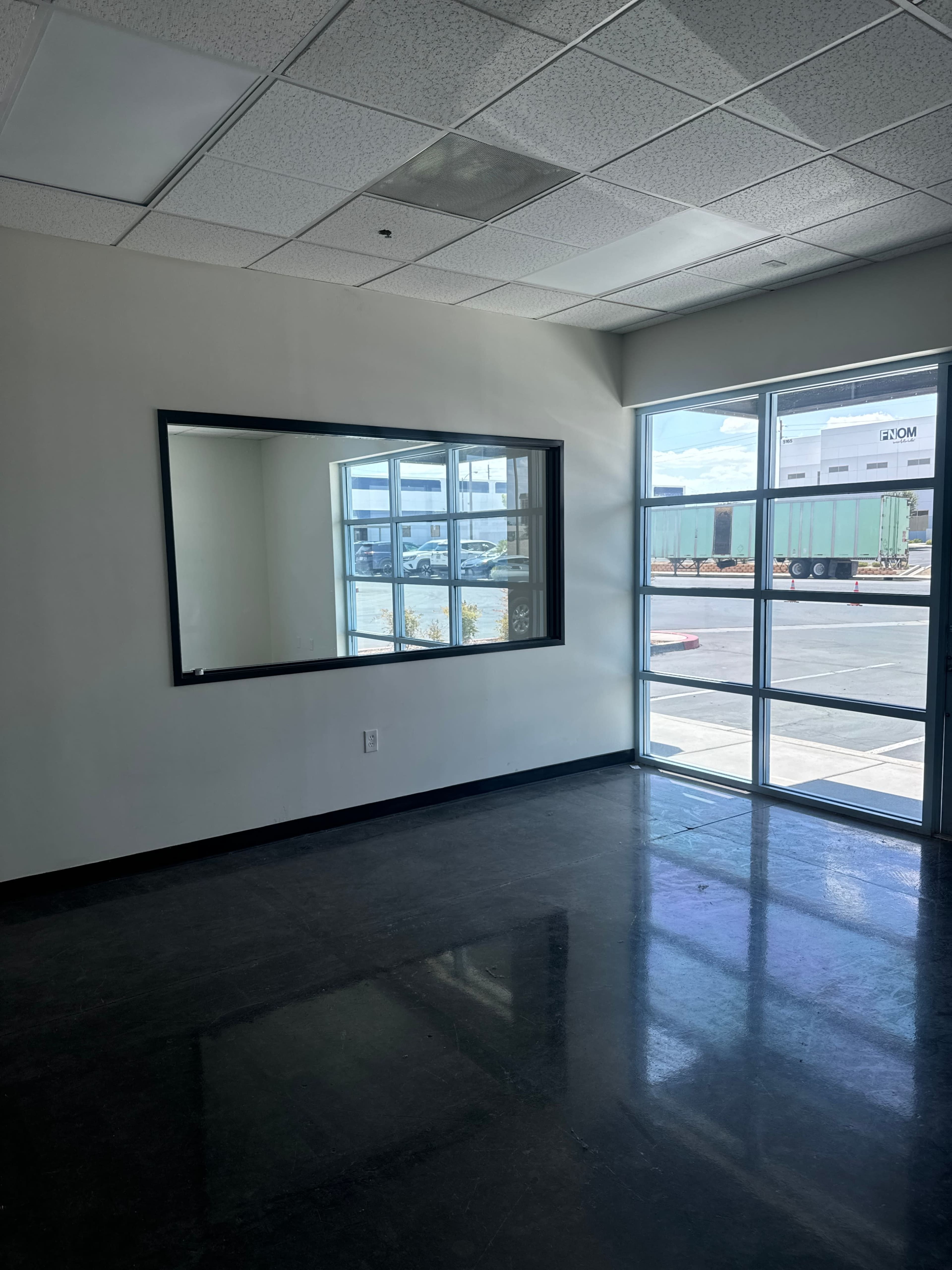 The image shows an empty room with a large mirror on one wall and a window that looks out onto a parking area with shipping containers.