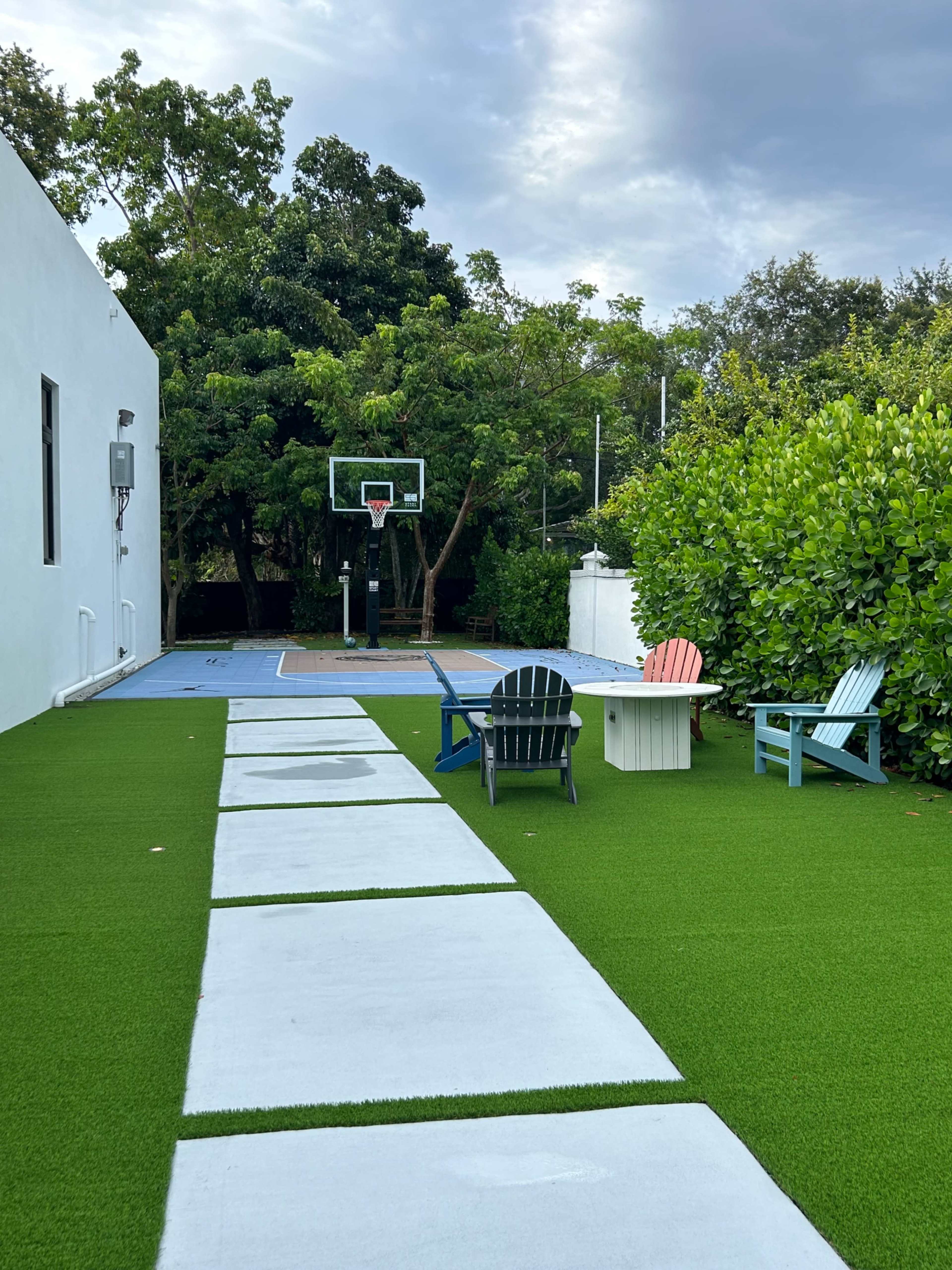 A pathway of stone slabs leads through a grassy area to a basketball court surrounded by lush greenery and colorful outdoor chairs.