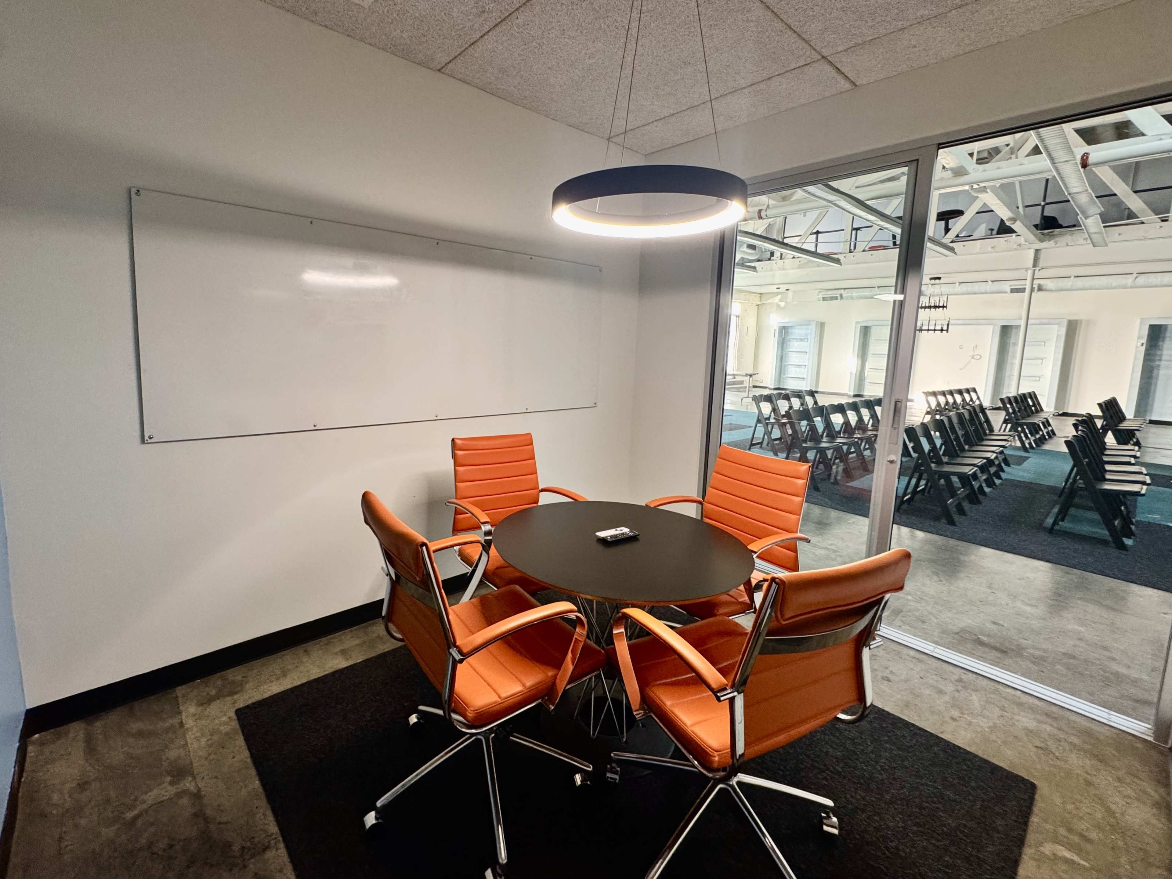 The image shows a small meeting room with a round black table, four orange chairs, a whiteboard on the wall, and a light fixture overhead.