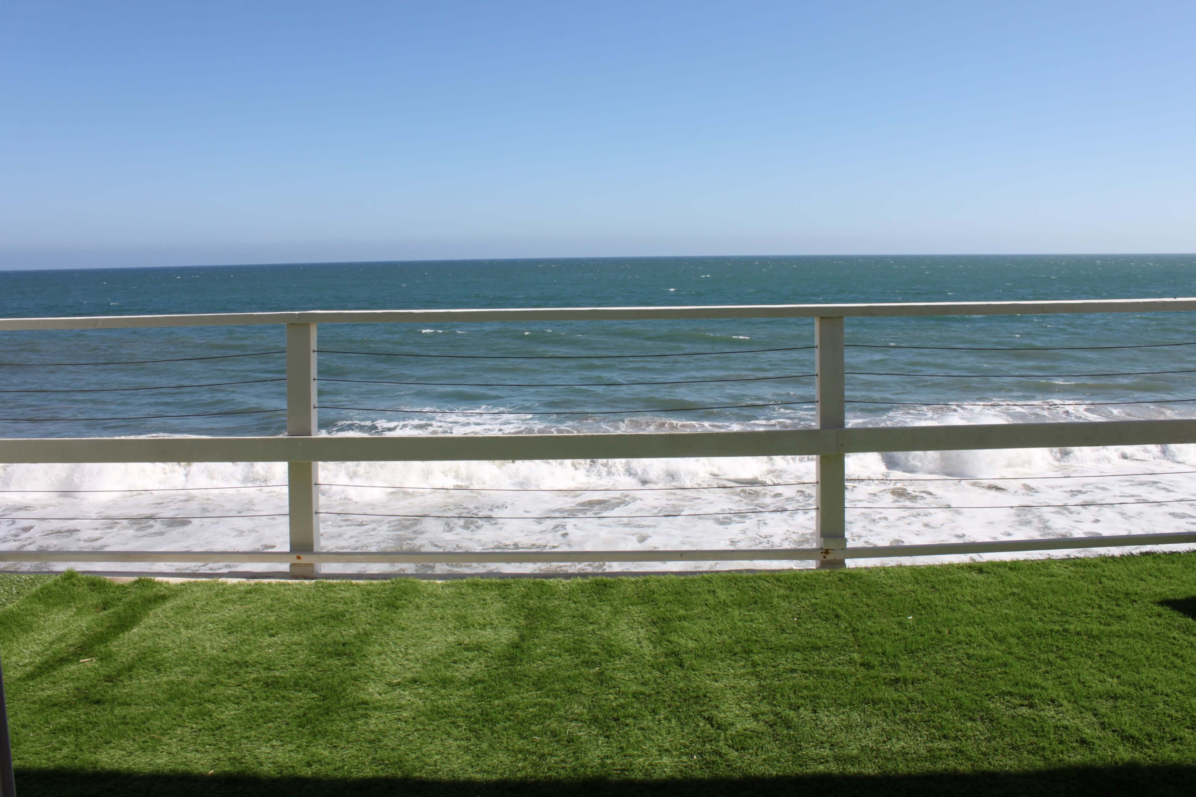 The image shows a view of a calm ocean with gentle waves rolling towards a grassy area, separated by a white railing.