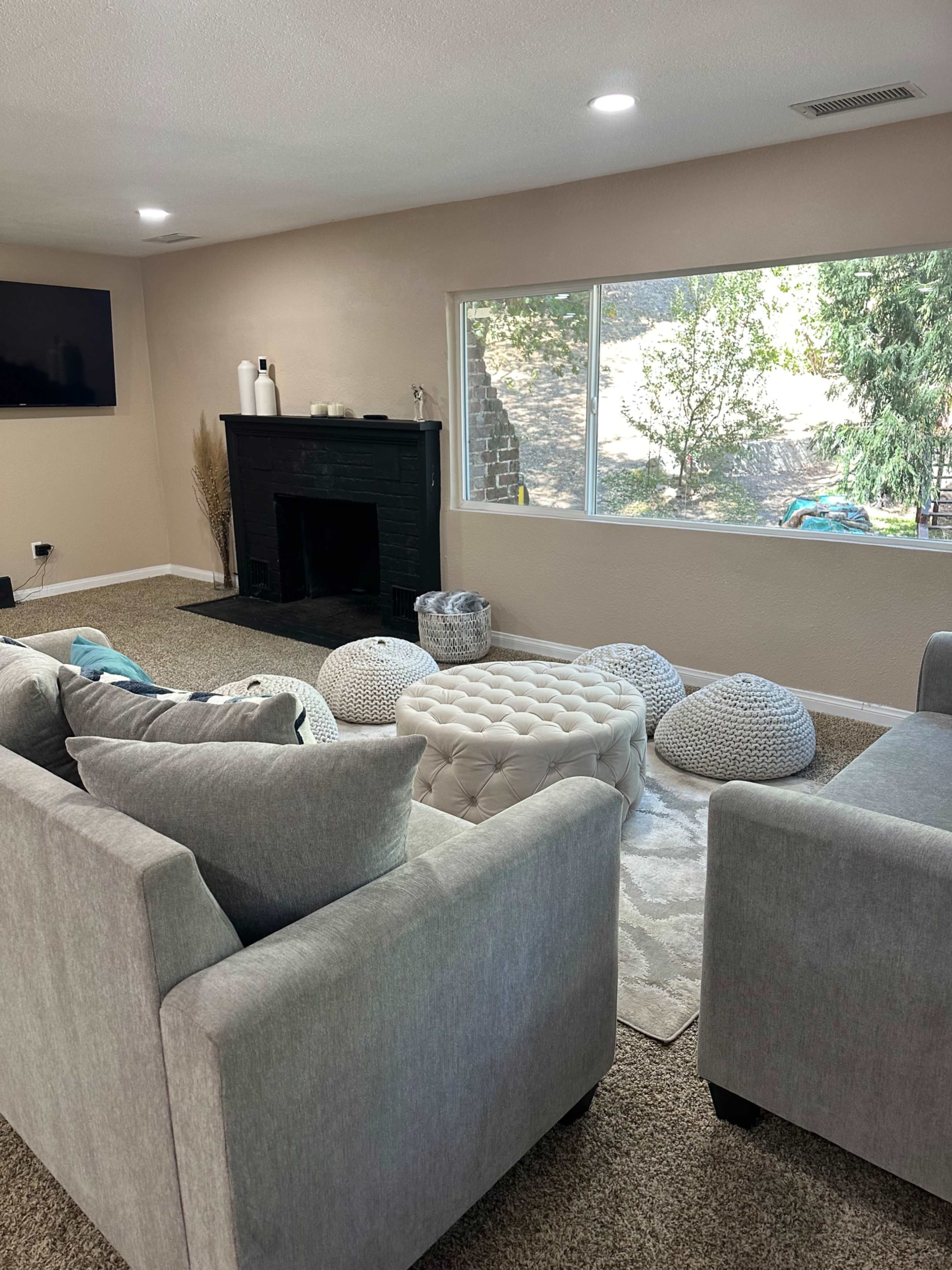 The living room features a gray sofa set, a tufted coffee table, and decorative poufs arranged around a black fireplace, with large windows offering a view of the outdoors.