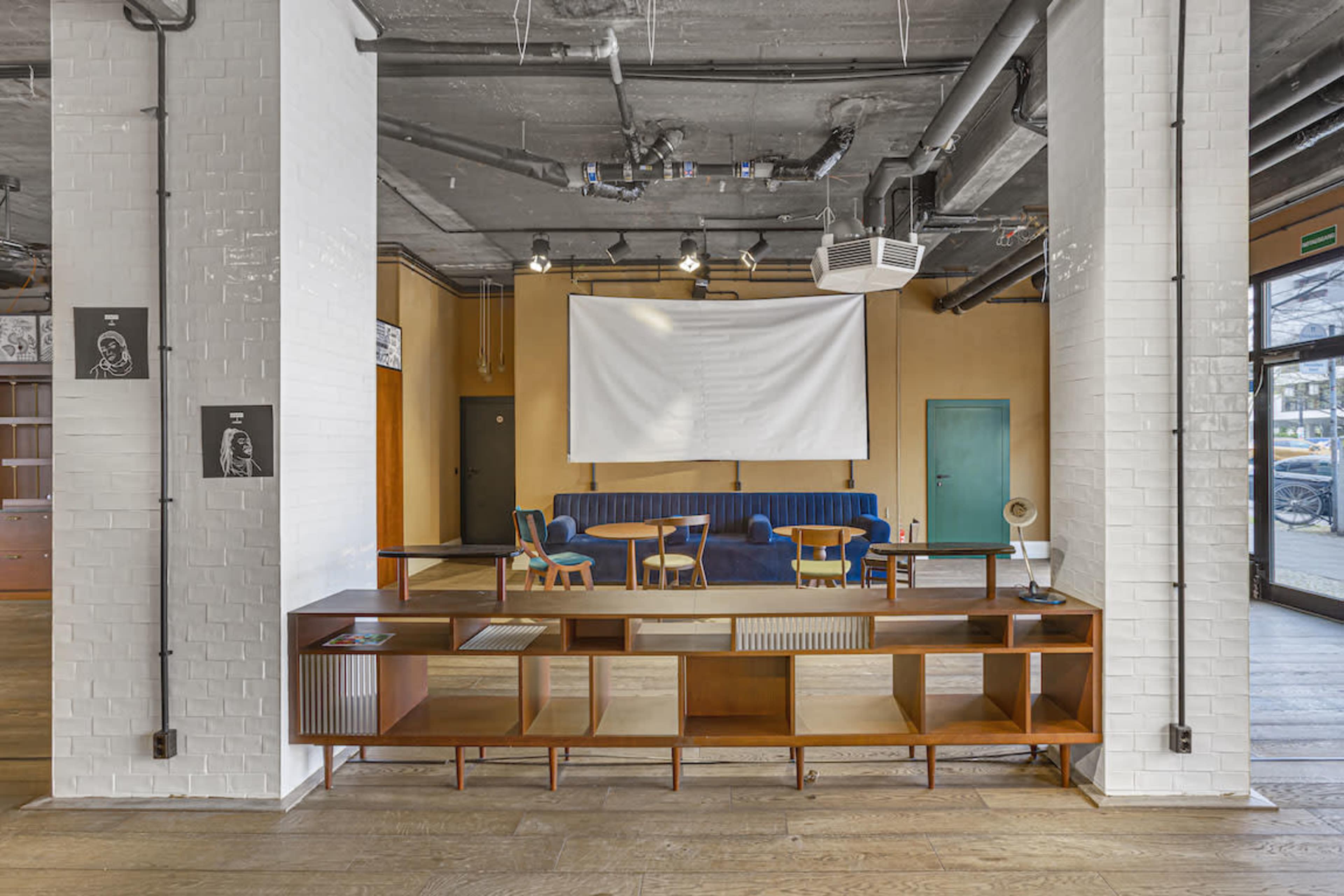 A modern interior space featuring a seating area with a blue couch, wooden tables, and chairs, alongside a large white projection screen and a wooden shelving unit.