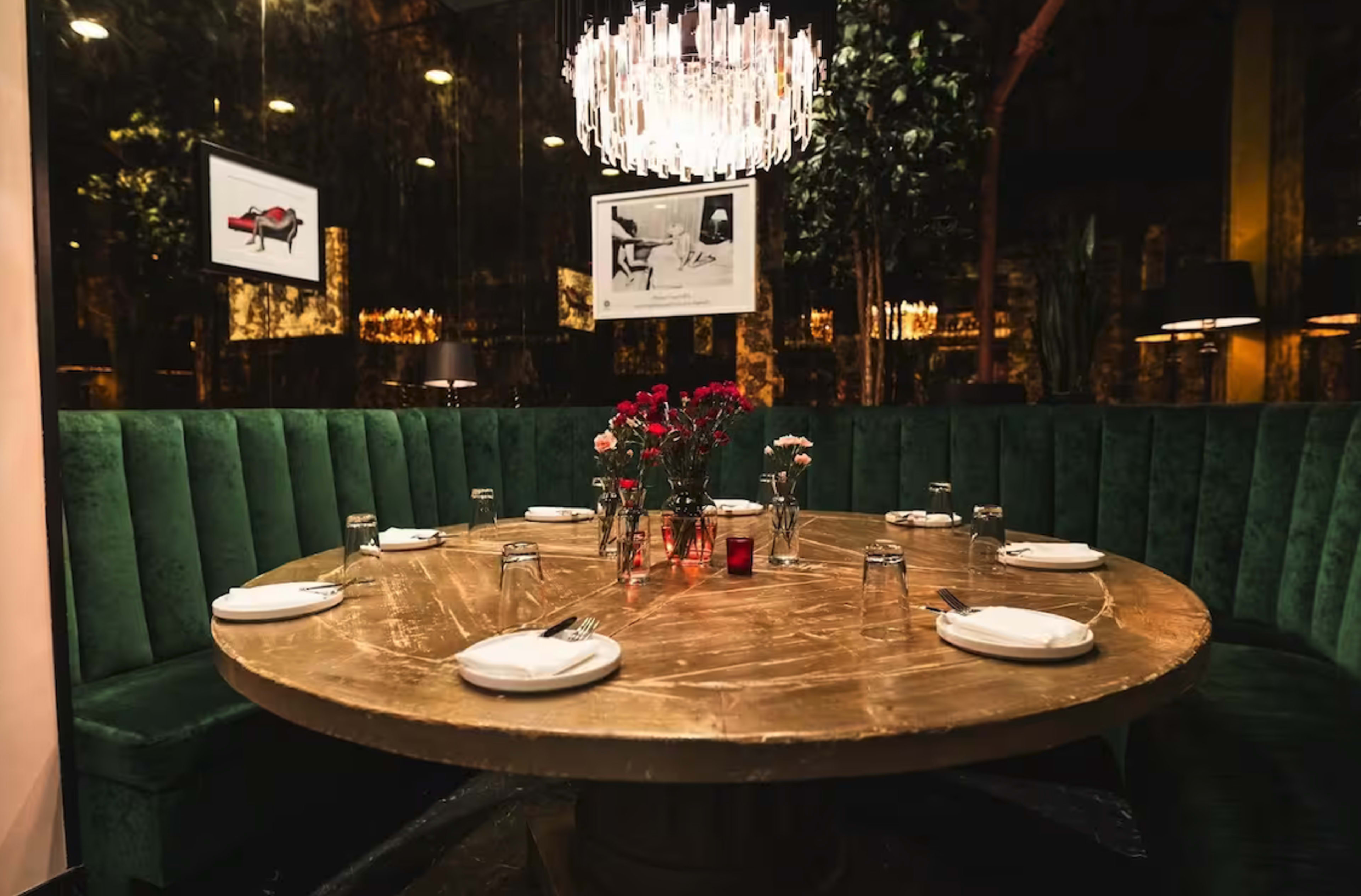 A round wooden table is set with plates and glasses, surrounded by green upholstered seating in a dimly lit restaurant.