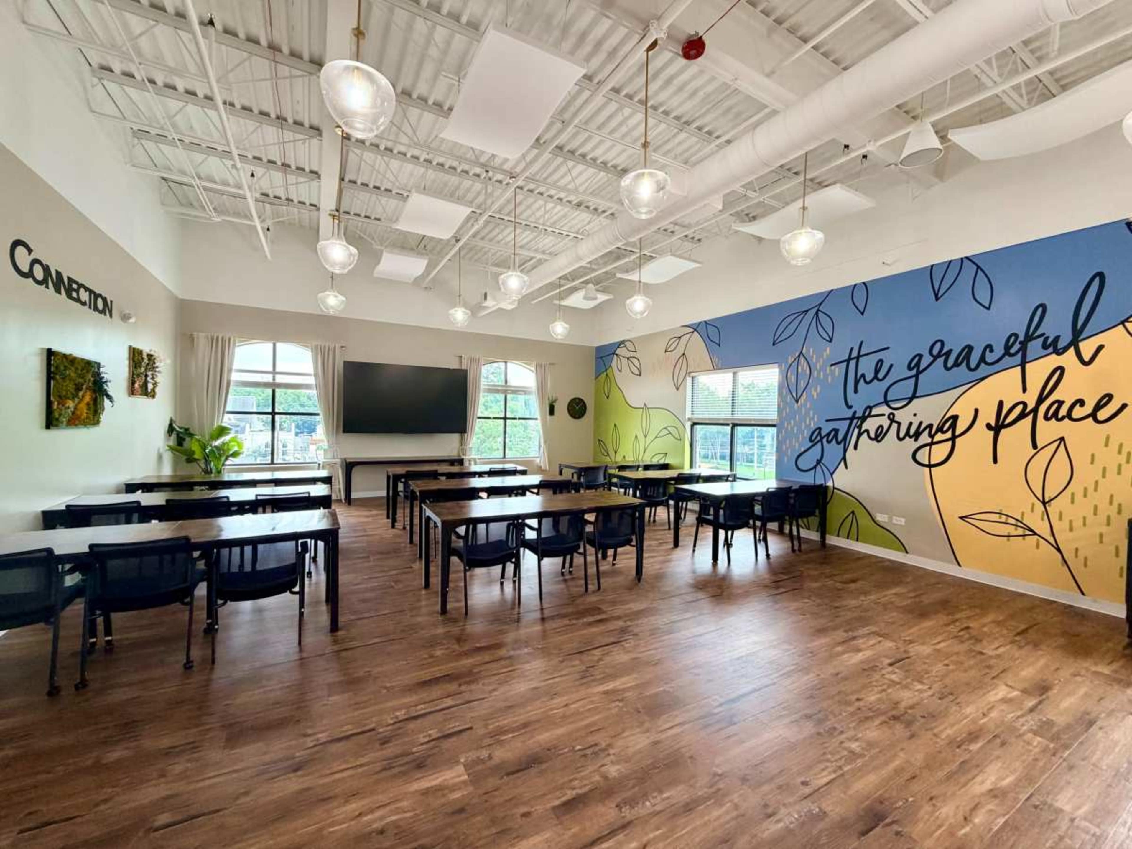 The image shows a well-lit meeting room with multiple tables and chairs arranged, featuring a colorful mural on one wall, and large windows that allow natural light to enter.
