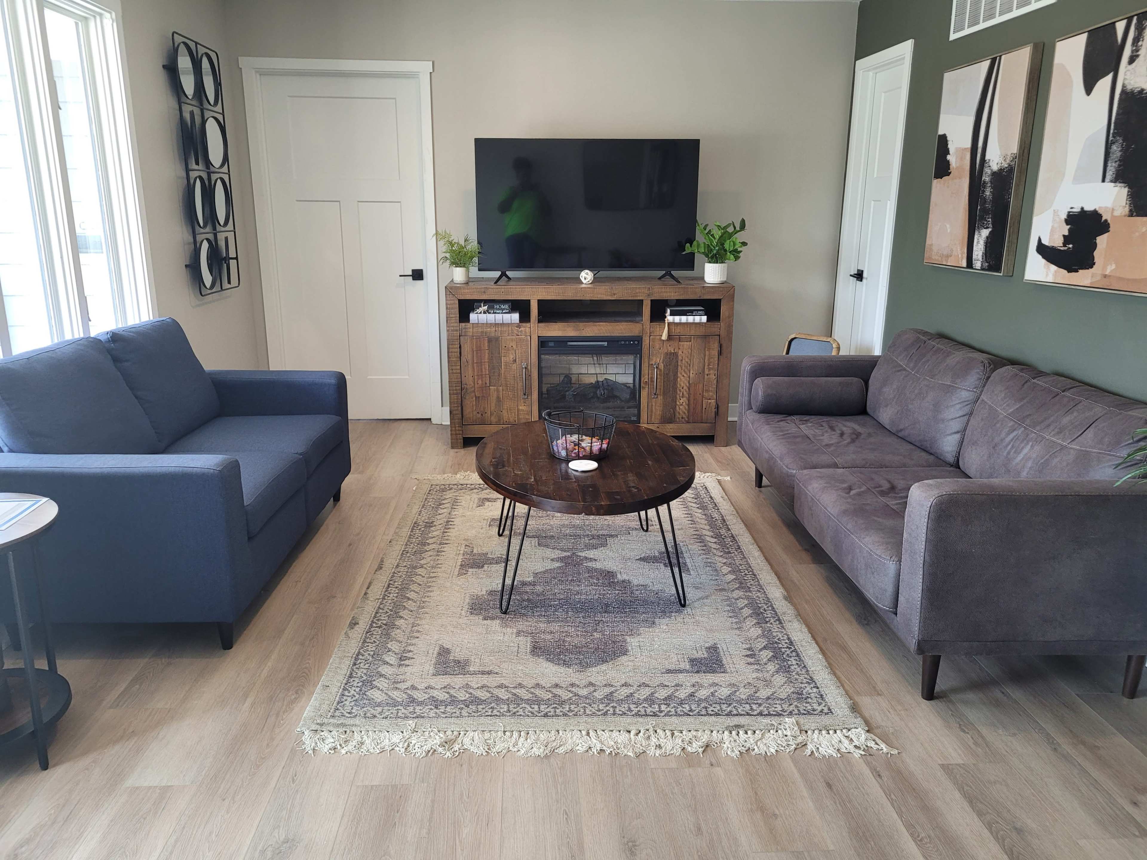 The living room features two sofas facing a wooden TV stand with a television, surrounded by a round coffee table and decorative items on a patterned area rug.