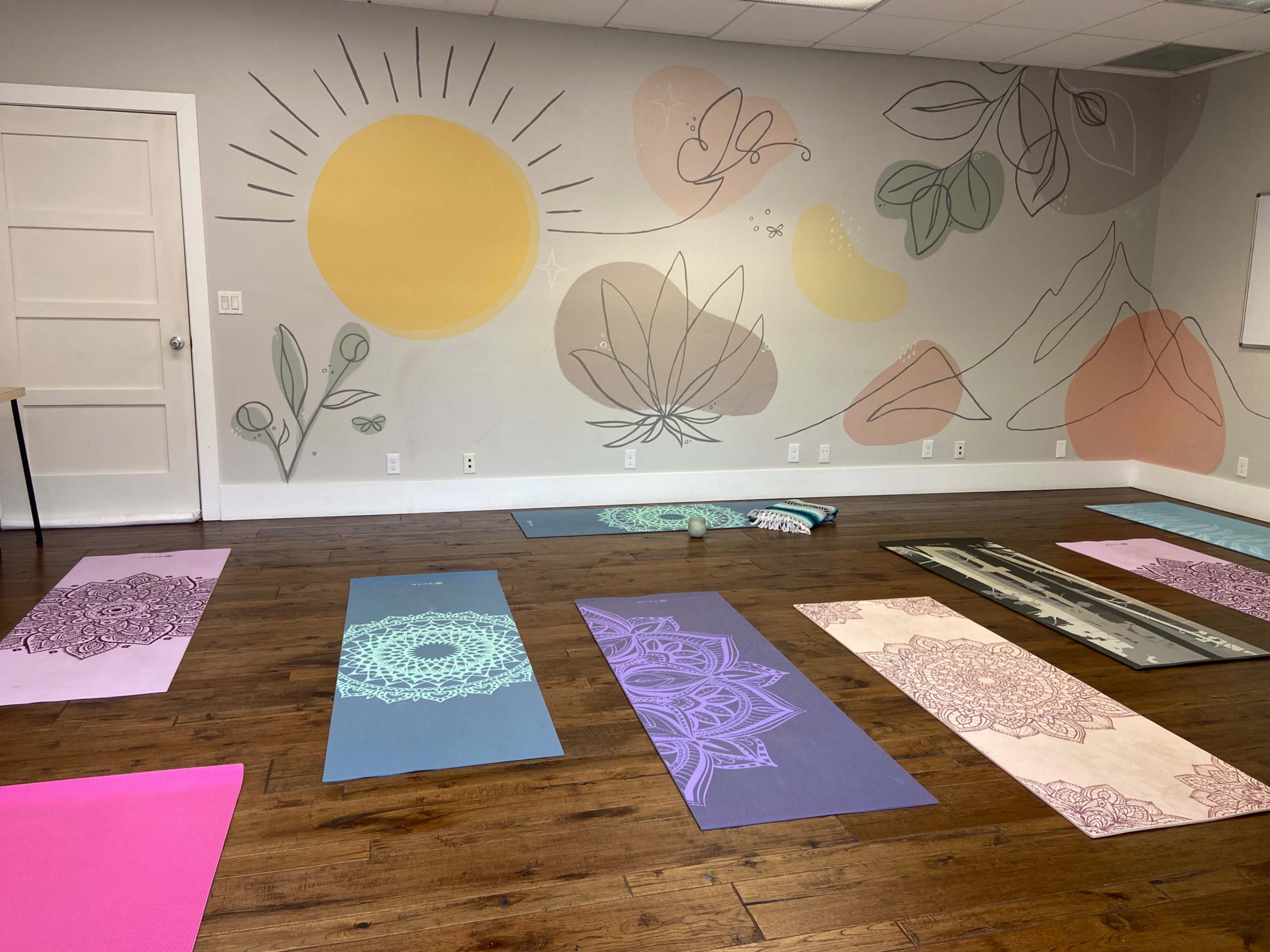 The image shows a yoga studio with several colorful mats arranged on a wooden floor beneath a wall adorned with a mural featuring a sun, leaves, and mountains.
