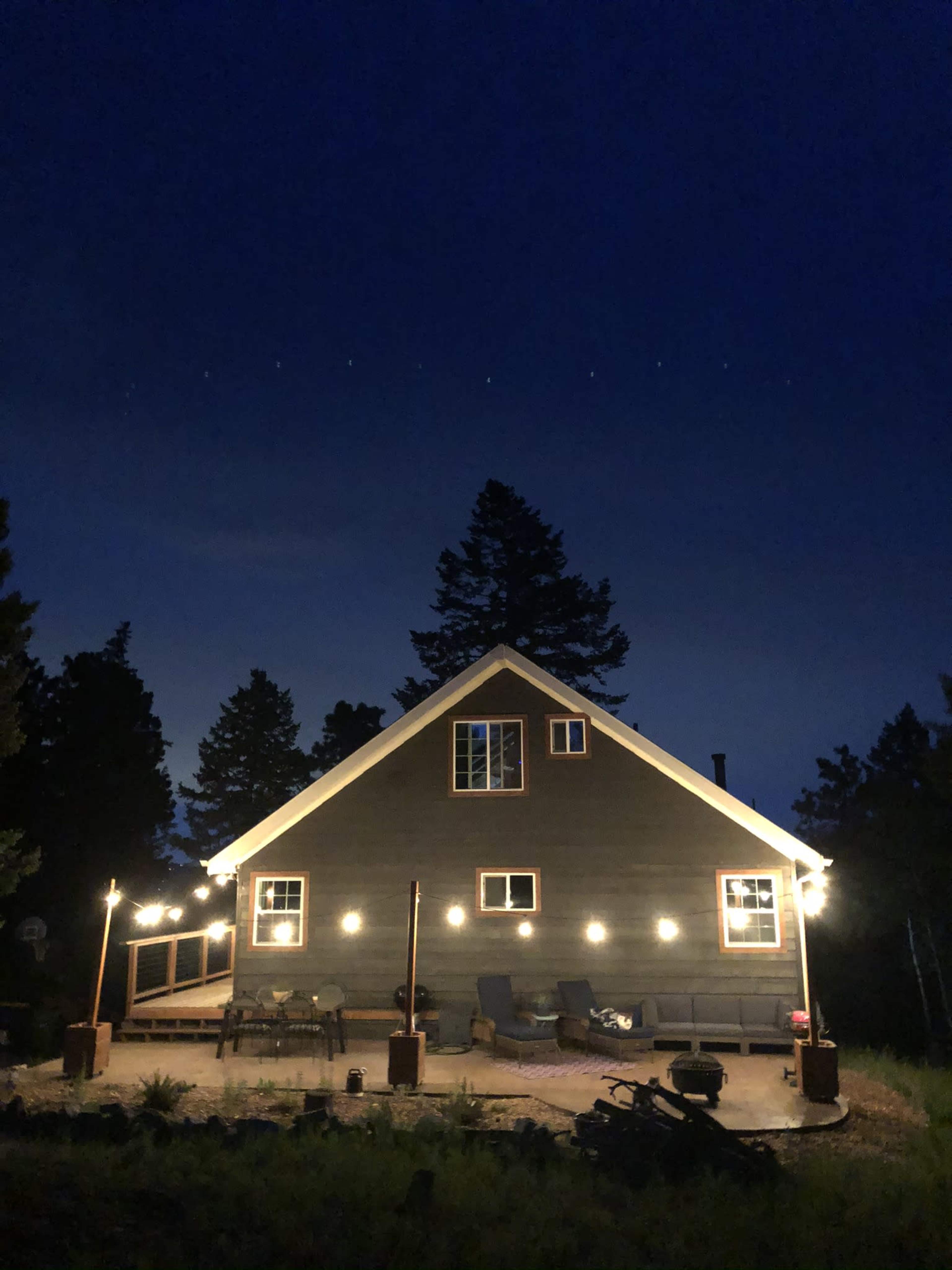 A dimly lit house with string lights illuminates its exterior against a dark night sky, surrounded by trees.