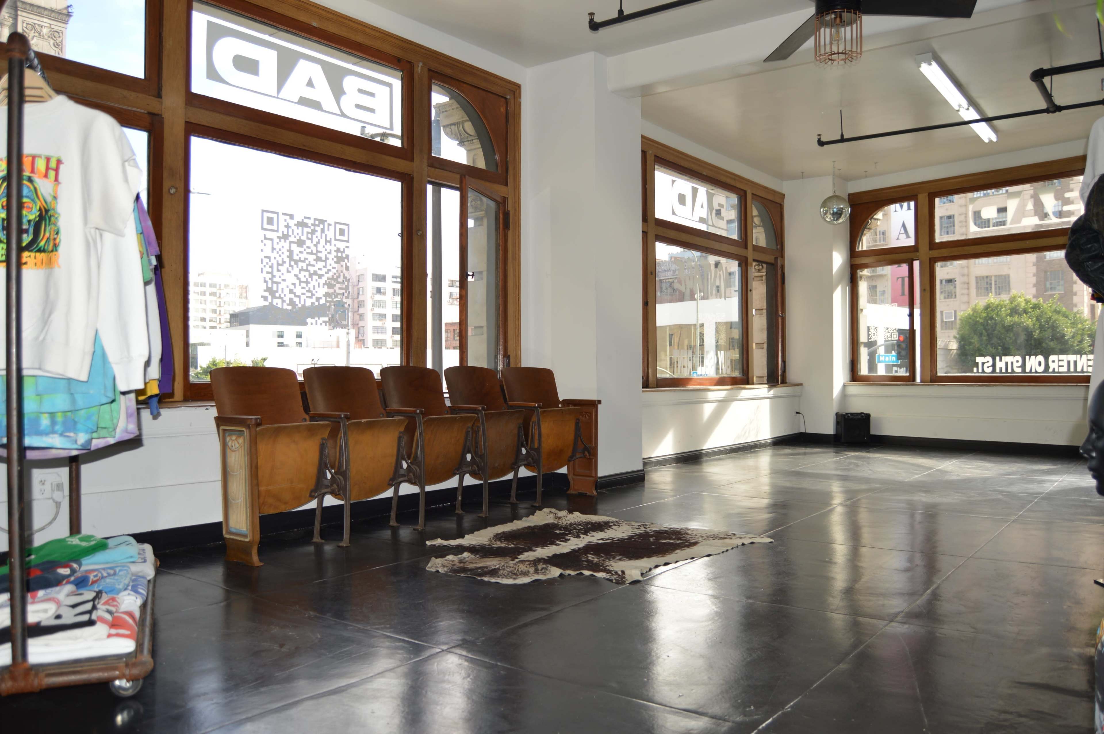 The image shows a spacious interior with wooden chairs aligned against a wall, a cowhide rug on the floor, and large windows displaying a city view.