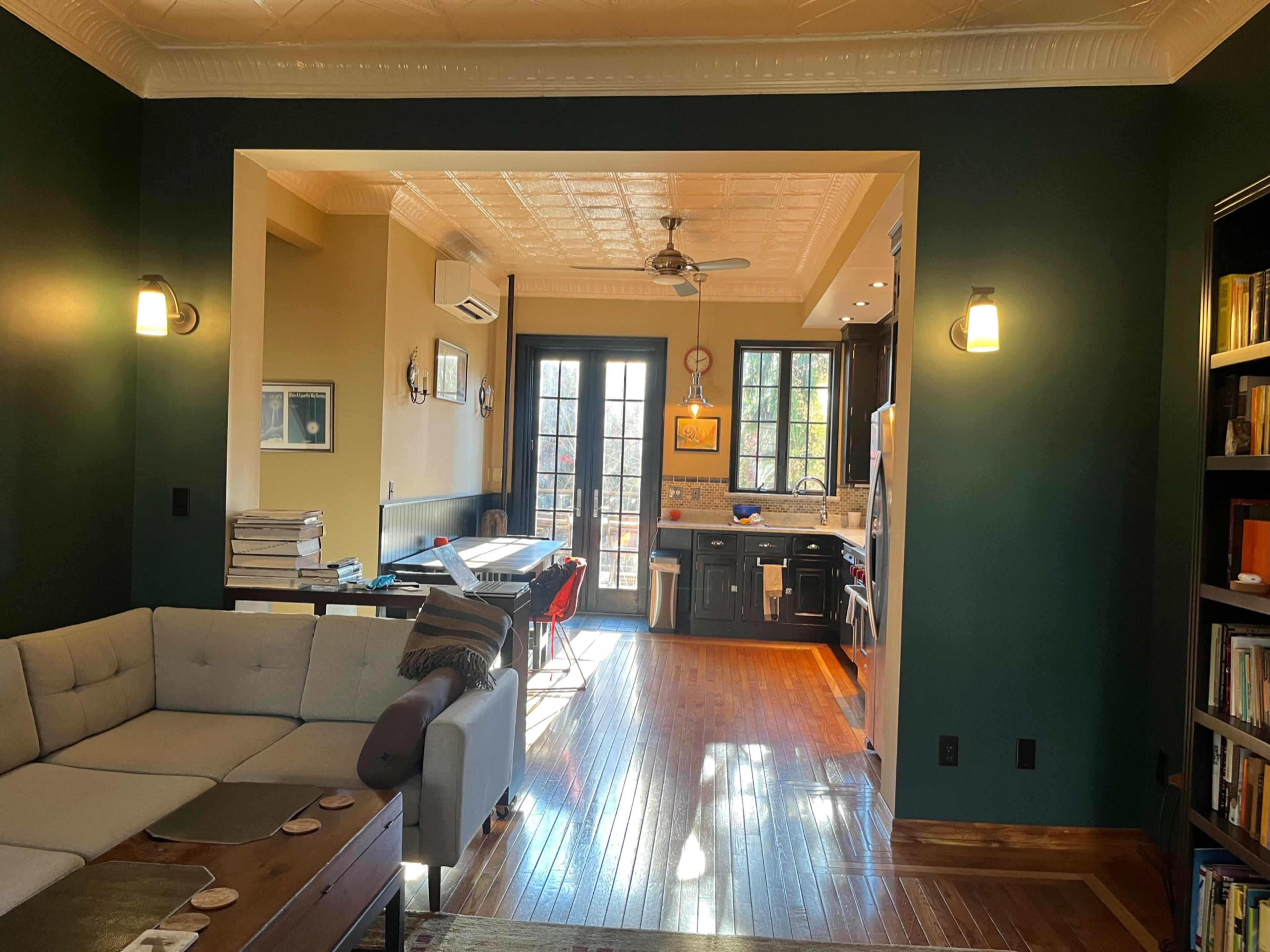 The image shows a living room with a beige sectional sofa, leading to an open kitchen area with dark wood floors and large windows.