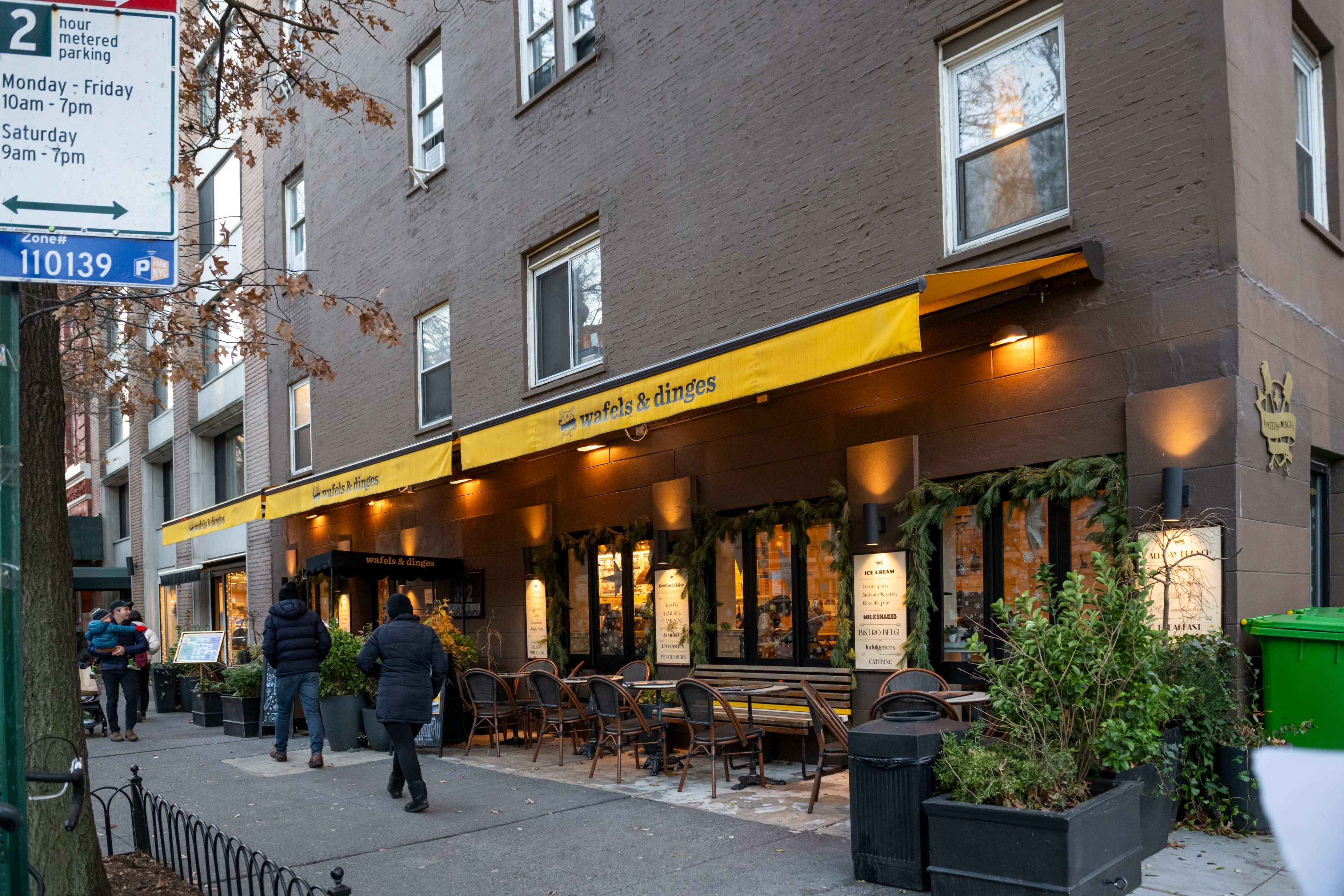 A restaurant with outdoor seating and yellow awnings is situated along a city street, with pedestrians walking by.