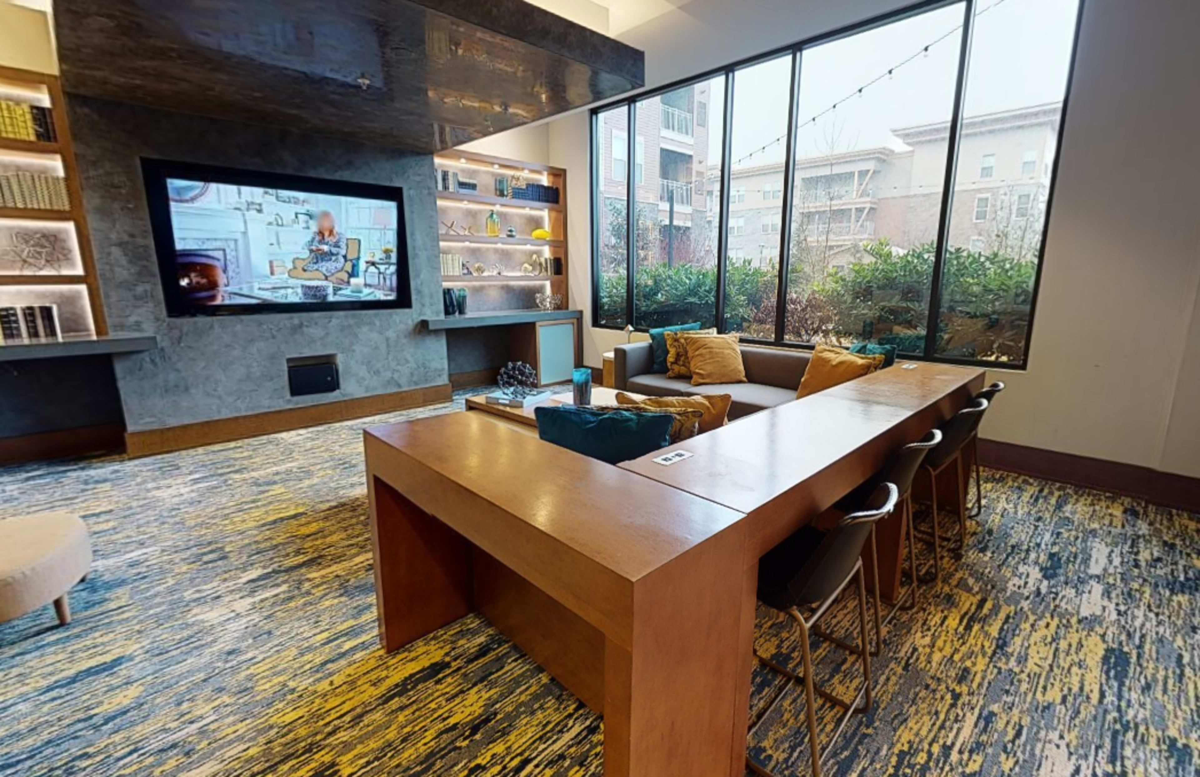 A modern lounge area with a large wall-mounted television, a wooden table surrounded by seating, and large windows displaying an outdoor view.