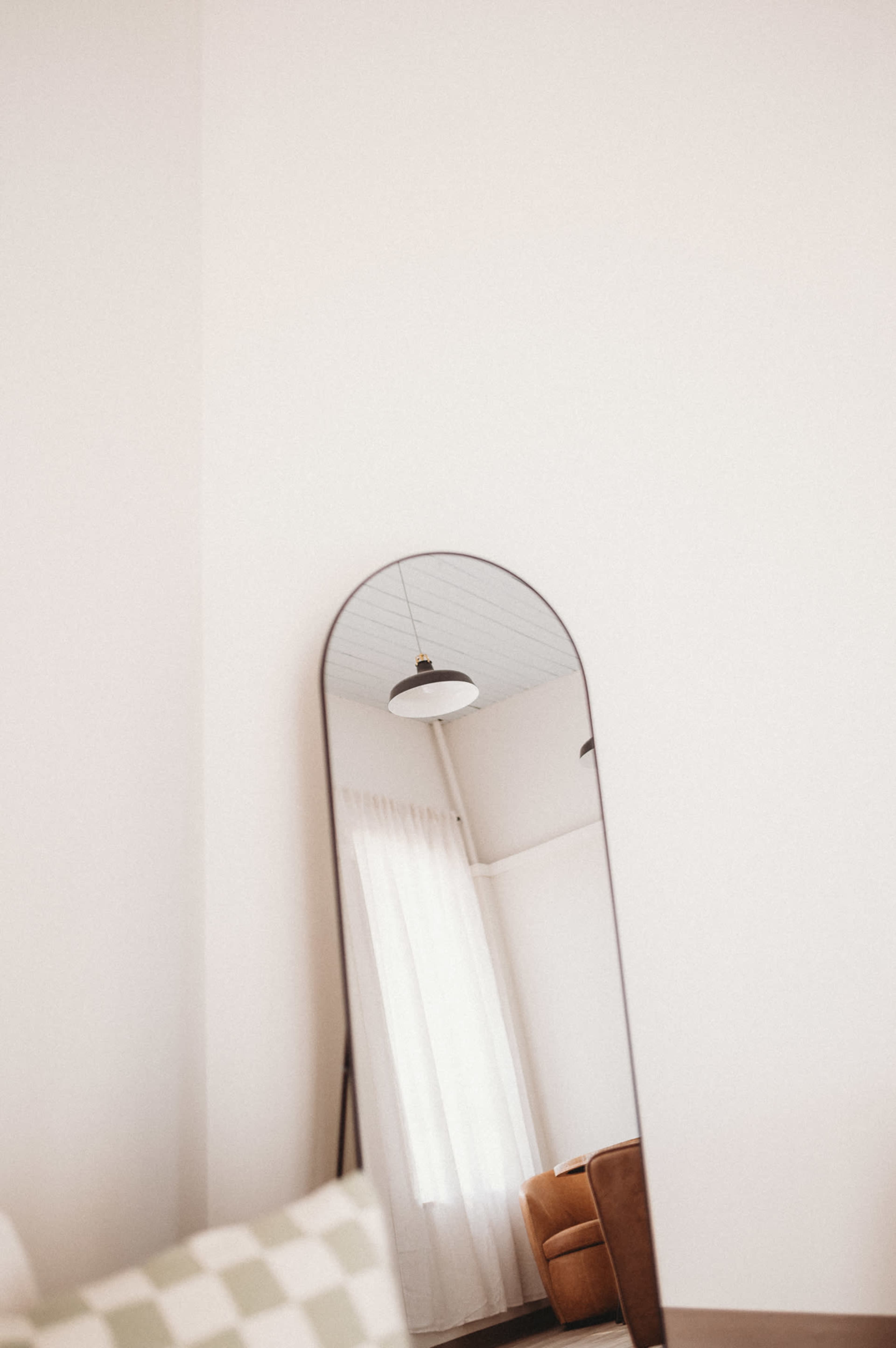 A tall, arched mirror reflects a room with a ceiling light and a curtain, beside a brown armchair.