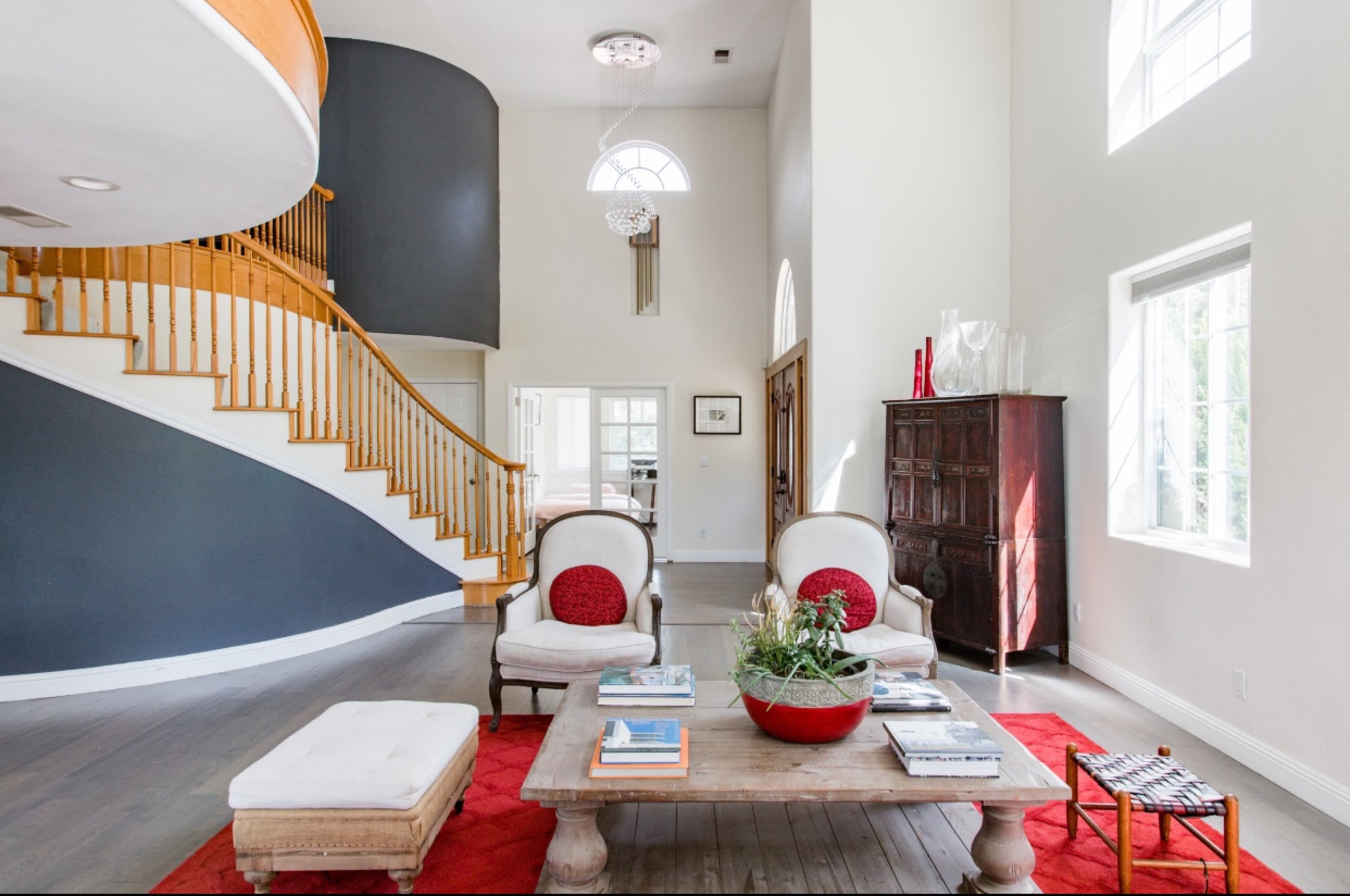 A spacious living room featuring a staircase with wood railings, two armchairs with red pillows, a wooden coffee table, and large windows that allow natural light to enter.