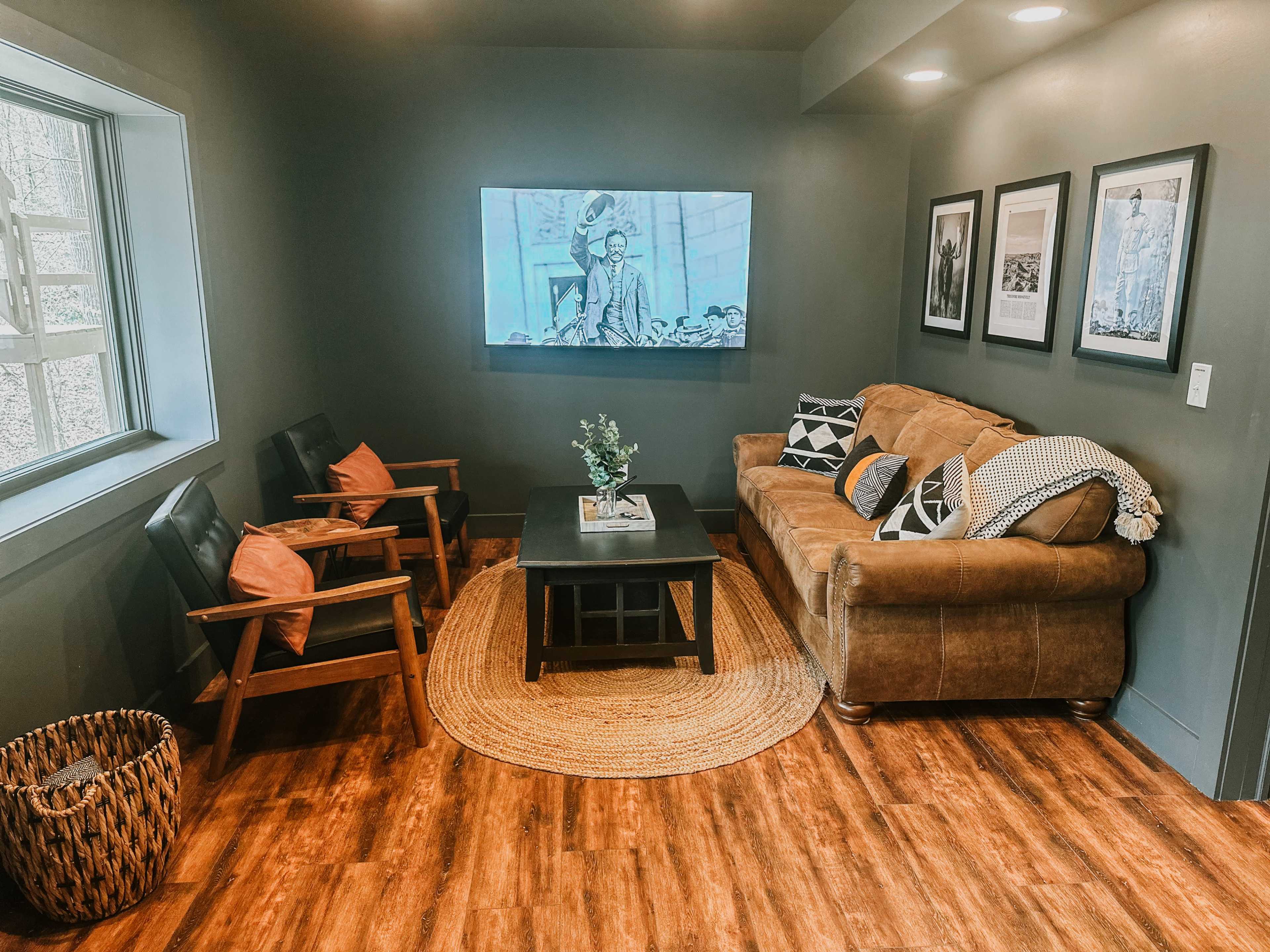 A cozy living area features a brown sofa, two wooden chairs, a coffee table, and a television mounted on the wall, all set against dark green walls and warm wood flooring.