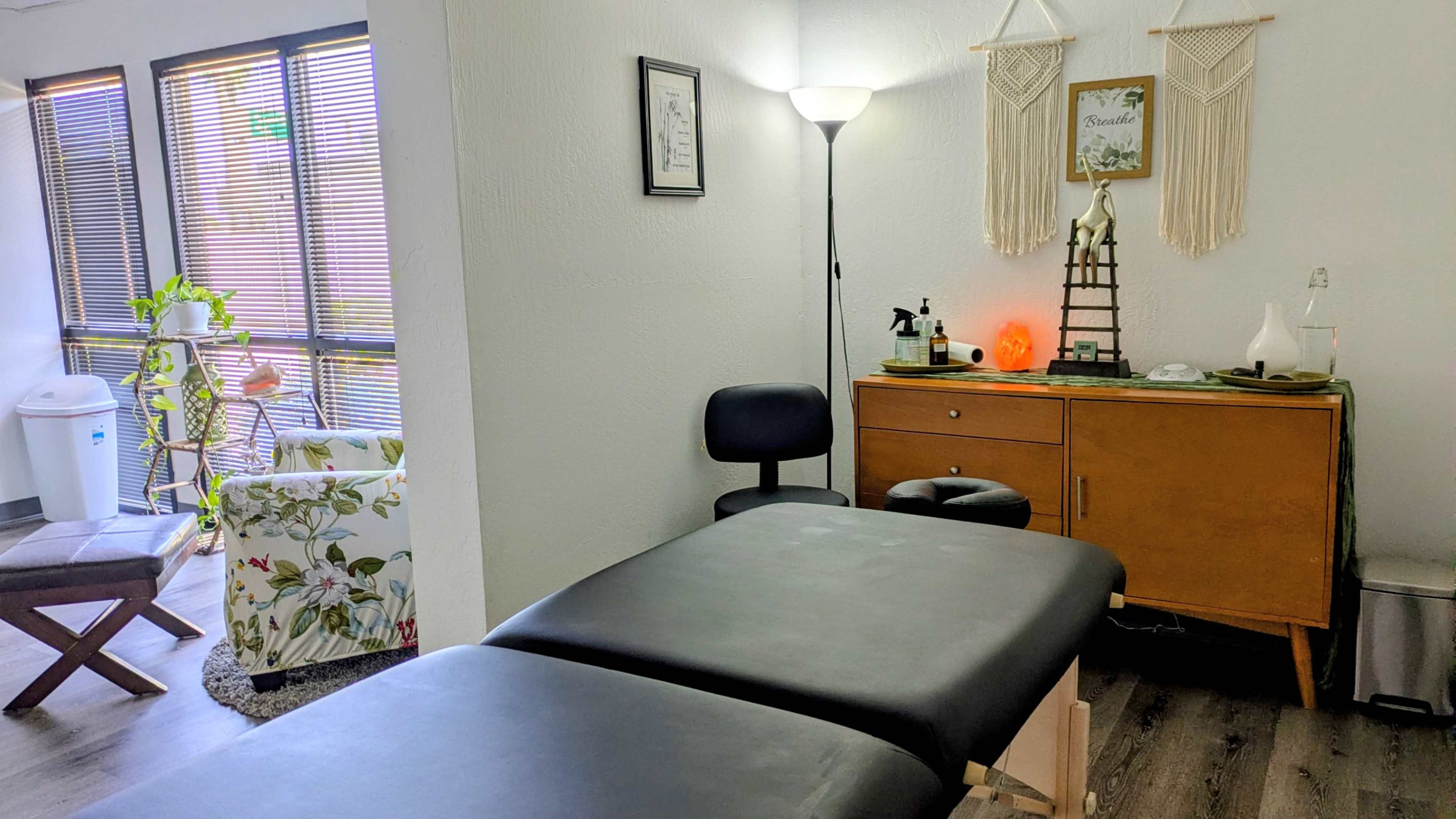 The image shows a treatment room with a massage table, a lamp, a wooden cabinet, and a chair, alongside decorative elements like plants and wall hangings.