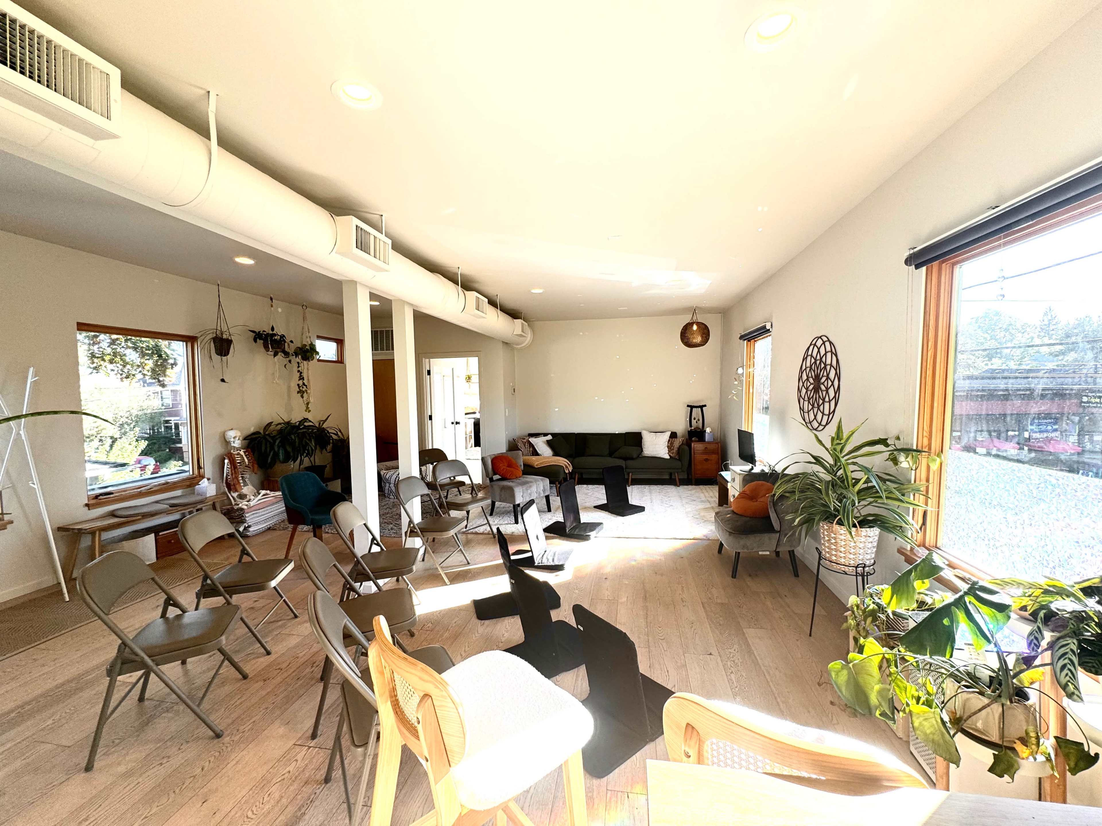 A bright room with large windows features a mix of seating including folding chairs and a sofa, surrounded by plants and minimal decor.