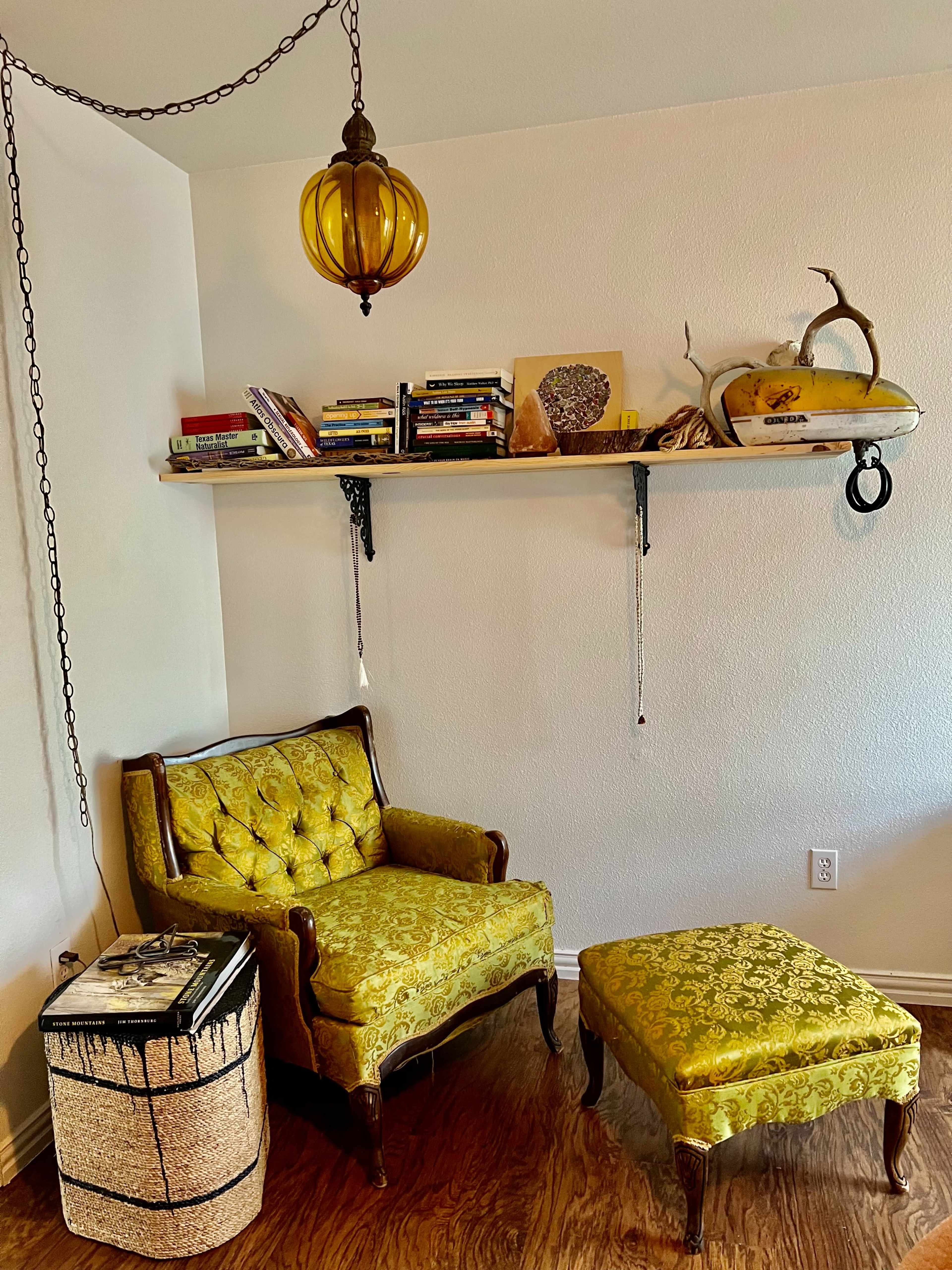 A cozy corner with a yellow tufted armchair, a matching ottoman, a shelf lined with books, and a pendant light hanging above.