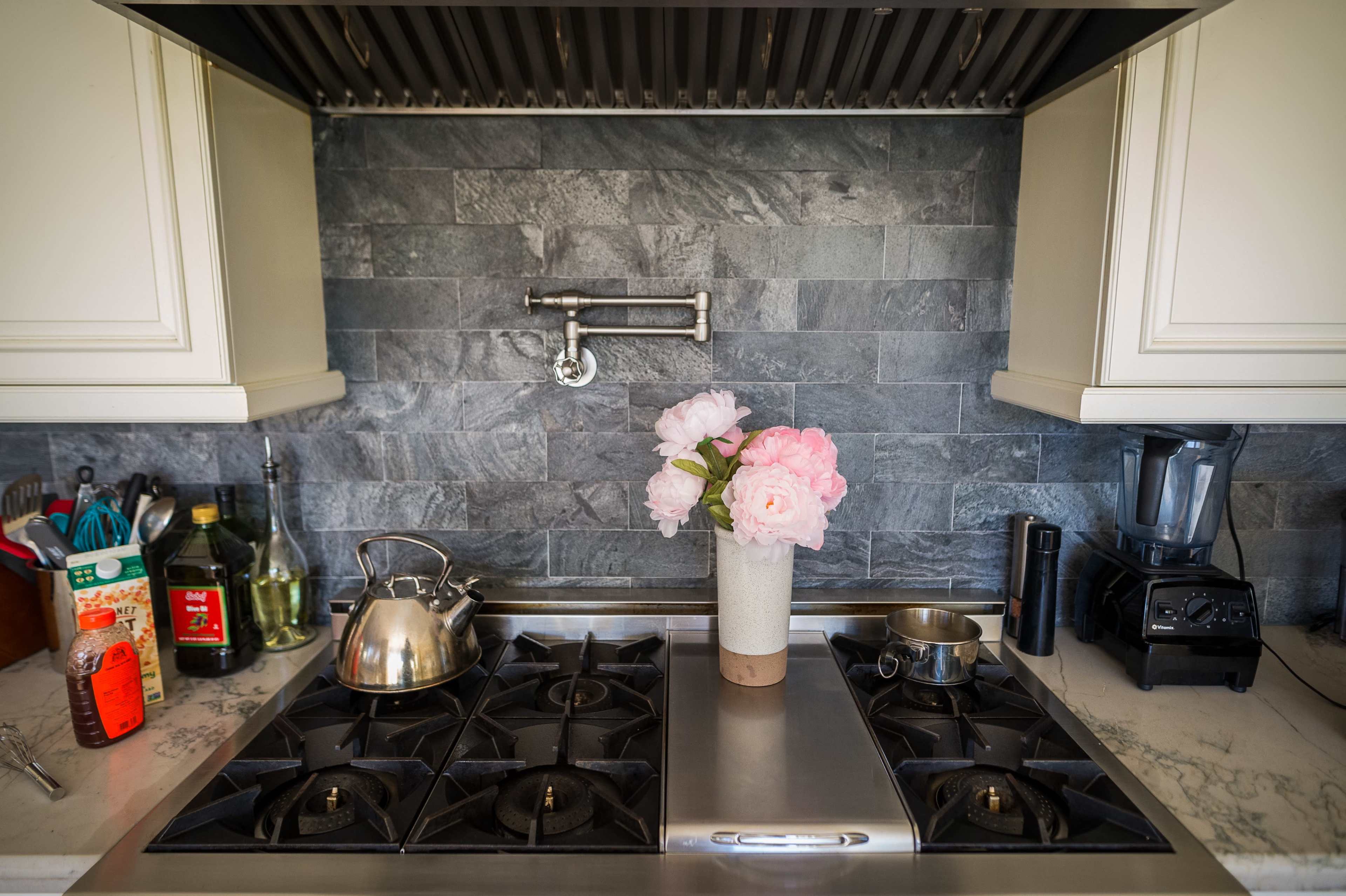 A modern kitchen countertop with a stainless steel gas stove, a kettle, a small pot, and a vase of pink flowers on a marble surface.