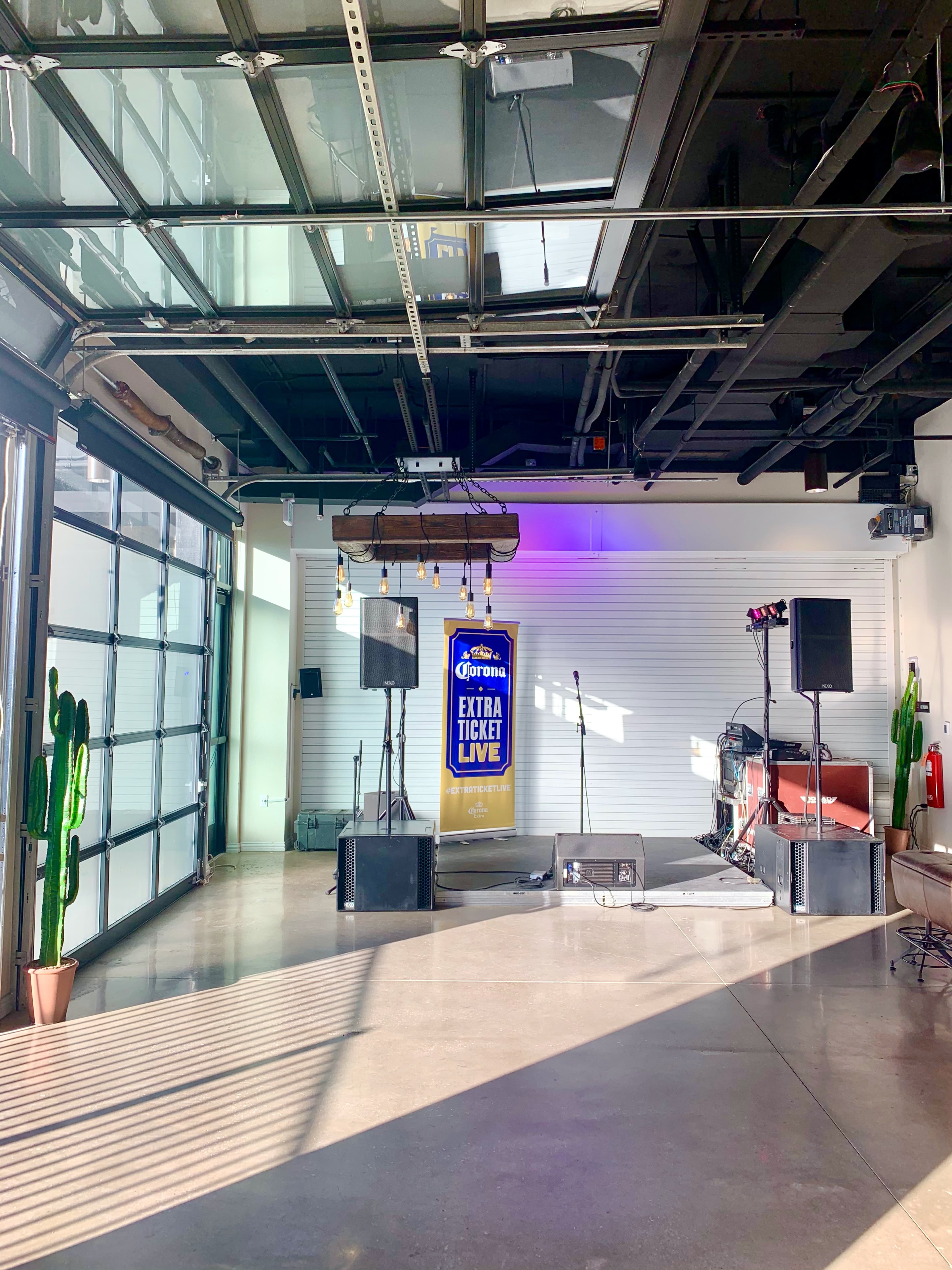 The scene shows a stage area in a modern indoor venue featuring a banner, microphones, and musical equipment, with large glass doors and potted cacti in the background.