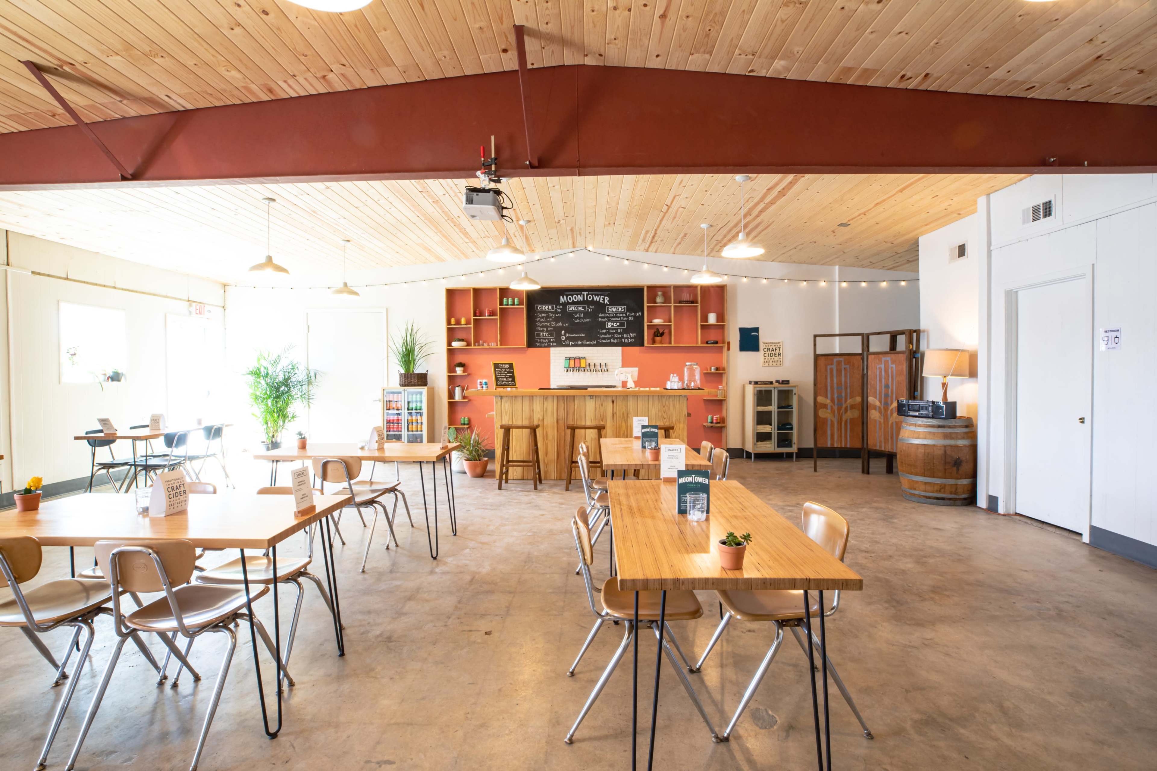 A spacious café interior with wooden tables, chairs, and a counter that features shelves and a drink dispenser against a backdrop of wooden beams and overhead lights.