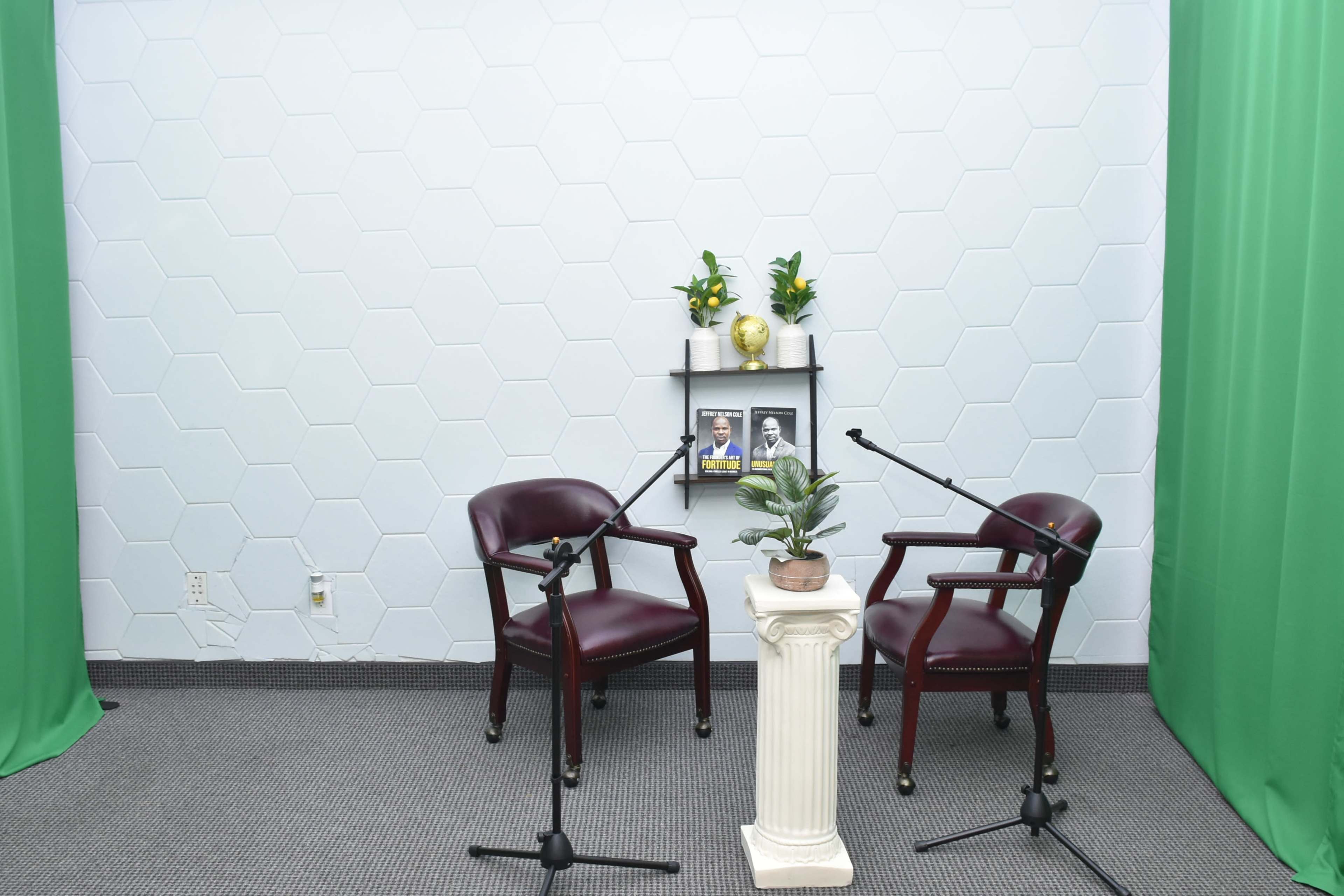The image shows two maroon chairs positioned facing a small table with a potted plant, framed photos, and microphones set on a patterned floor against a light-colored hexagonal wall with green curtains on either side.