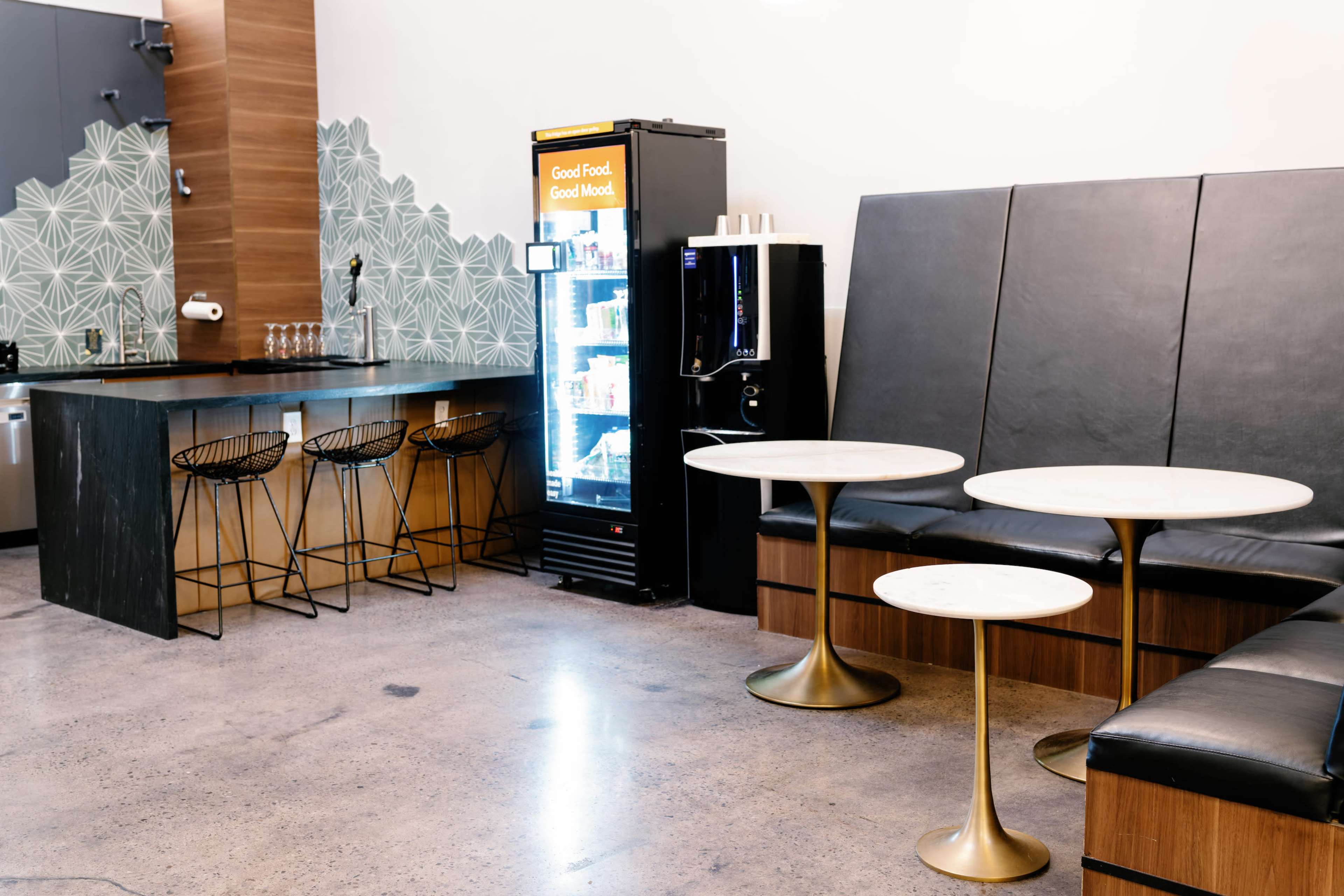 The image shows a modern café space featuring a beverage fridge, two small round tables with gold bases, black leather seating, and stylish bar stools at a counter.