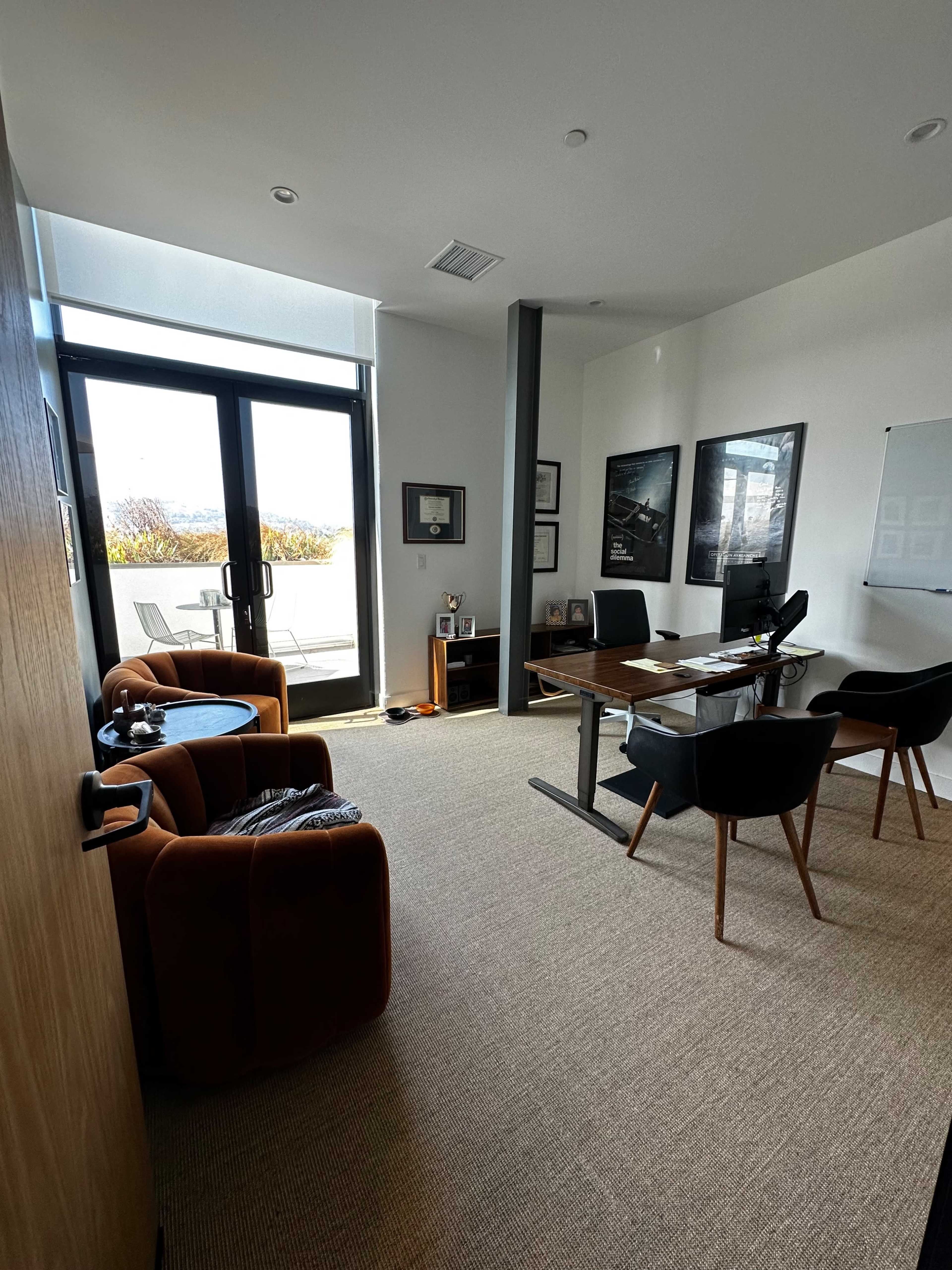 A modern office features a desk, chairs, and large windows leading to an outdoor area.